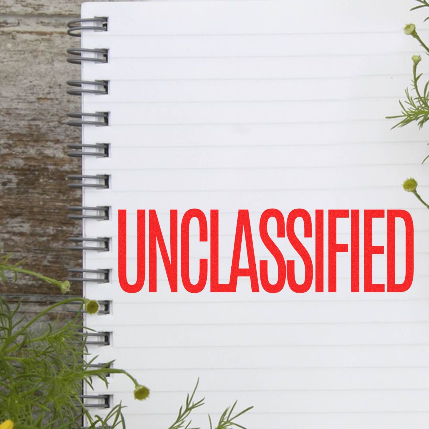 Large Pre-Inked Unclassified Stamp in red ink on a white spiral notebook, surrounded by green plants on a wooden surface.