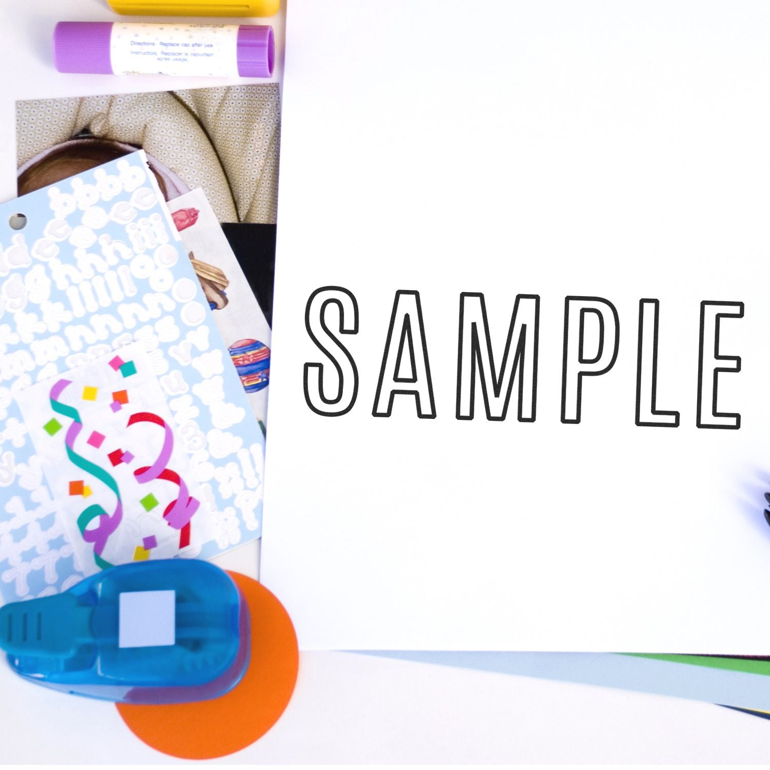 Large Pre-Inked Outline Sample Stamp on white paper, surrounded by colorful stationery items including stickers, glue stick, and a hole puncher.