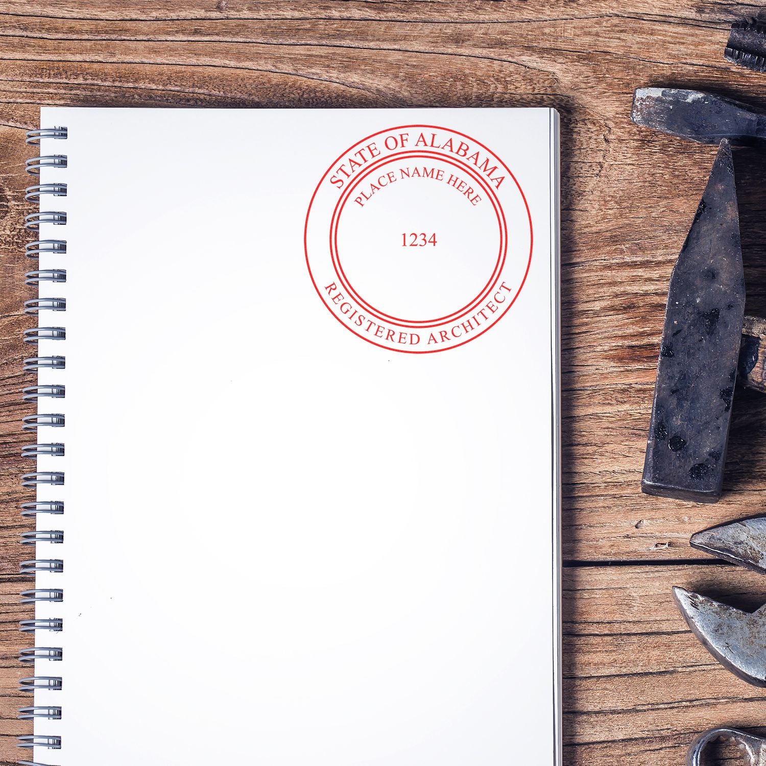 Self Inking Alabama Architect Stamp imprint on a blank spiral notebook, placed on a wooden desk with tools nearby.