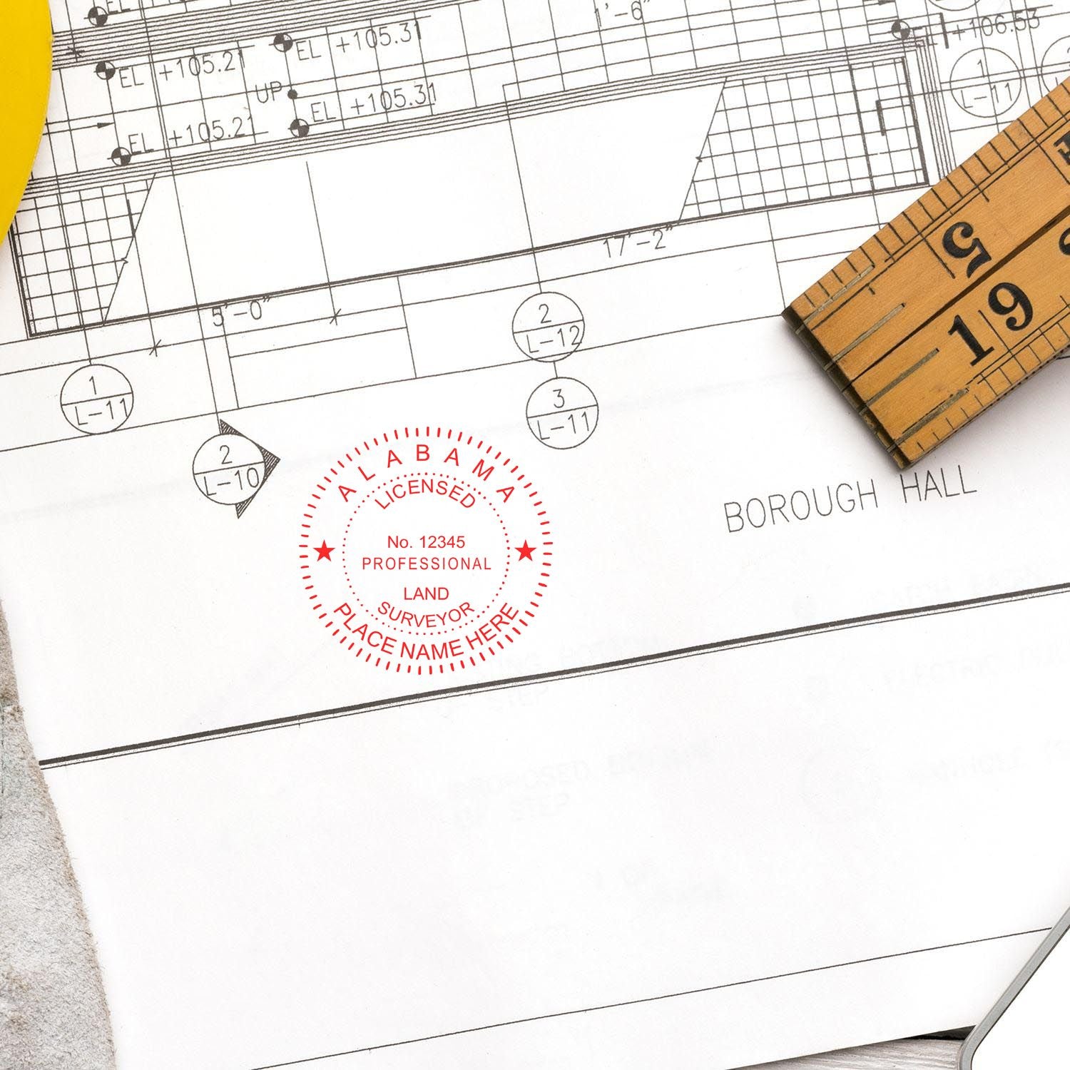 Self Inking Alabama Land Surveyor Stamp on architectural blueprint with a wooden ruler and yellow pen nearby.