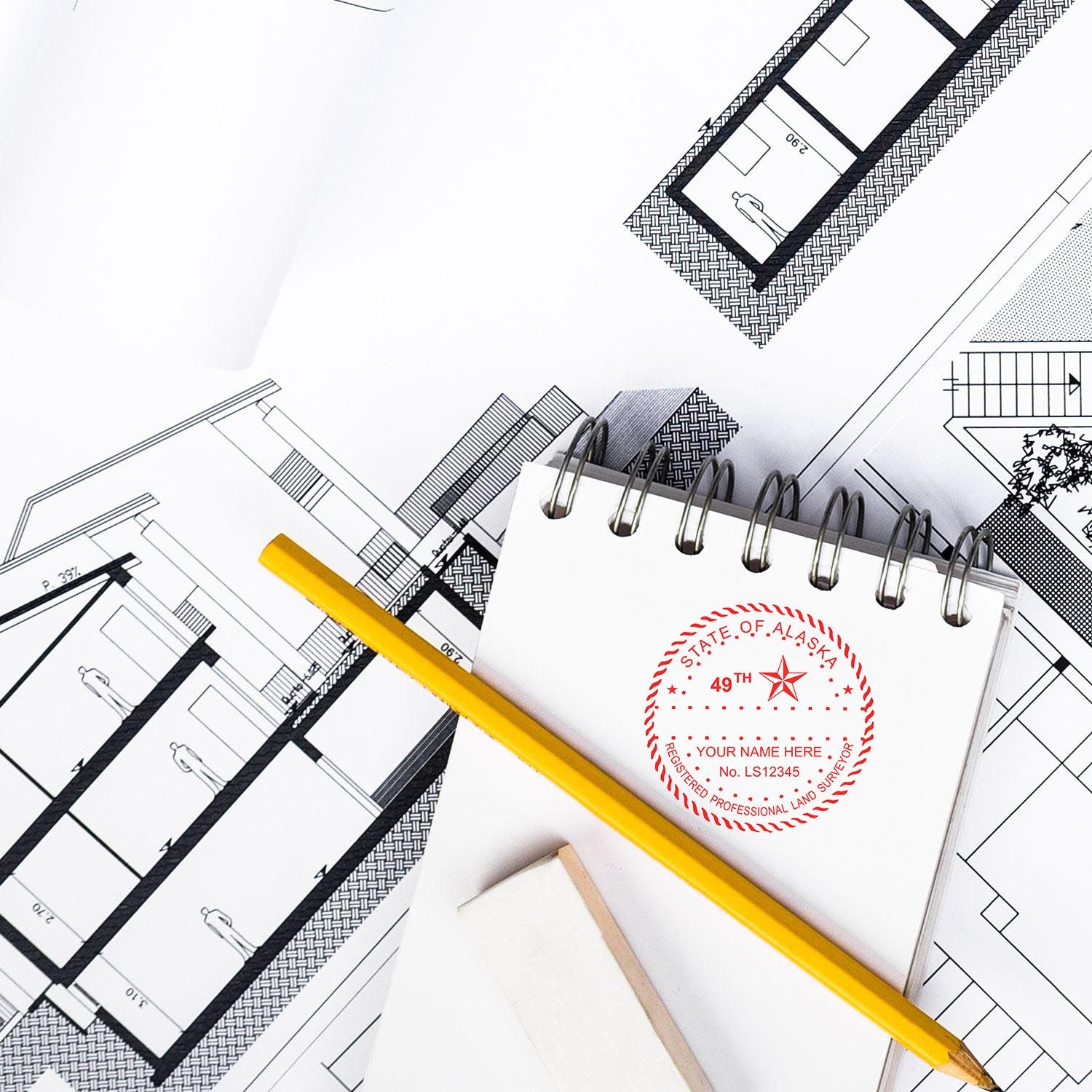 Self Inking Alaska Land Surveyor Stamp on a notepad, surrounded by architectural blueprints, a pencil, and an eraser.