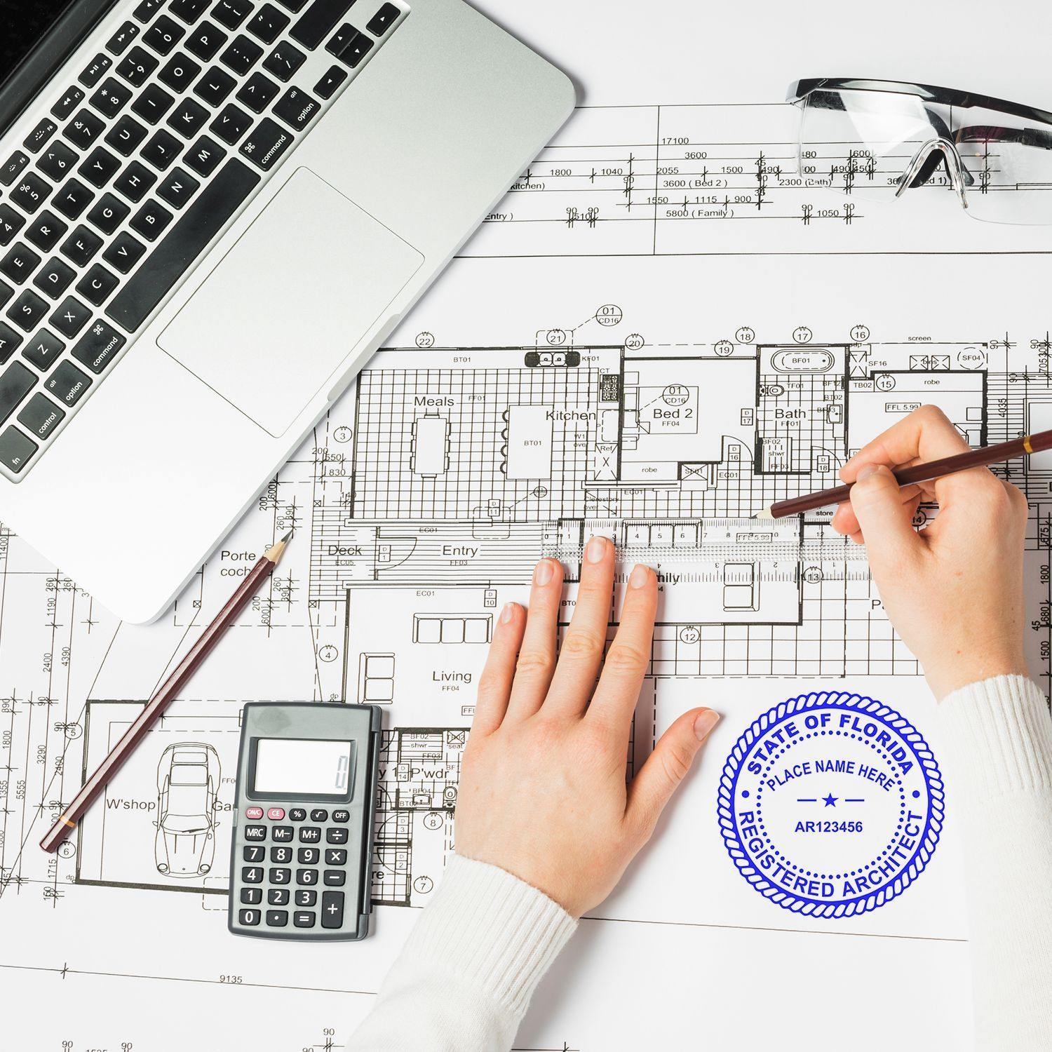 A hand using the Architect MaxLight Pre Inked Rubber Stamp of Seal on architectural blueprints, with a laptop, calculator, and glasses nearby.