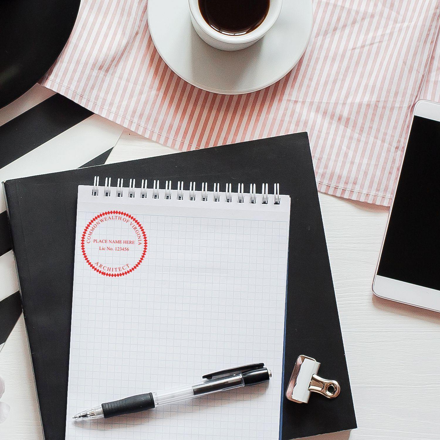 A desk with a notepad stamped with the Xstamper Architect Pre-Inked Rubber Stamp of Seal, a pen, a cup of coffee, a smartphone, and a clipboard.