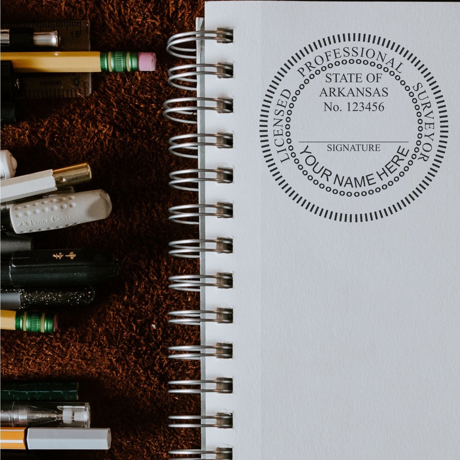 Self Inking Arkansas Land Surveyor Stamp on a spiral notebook, surrounded by pens and pencils on a brown surface.