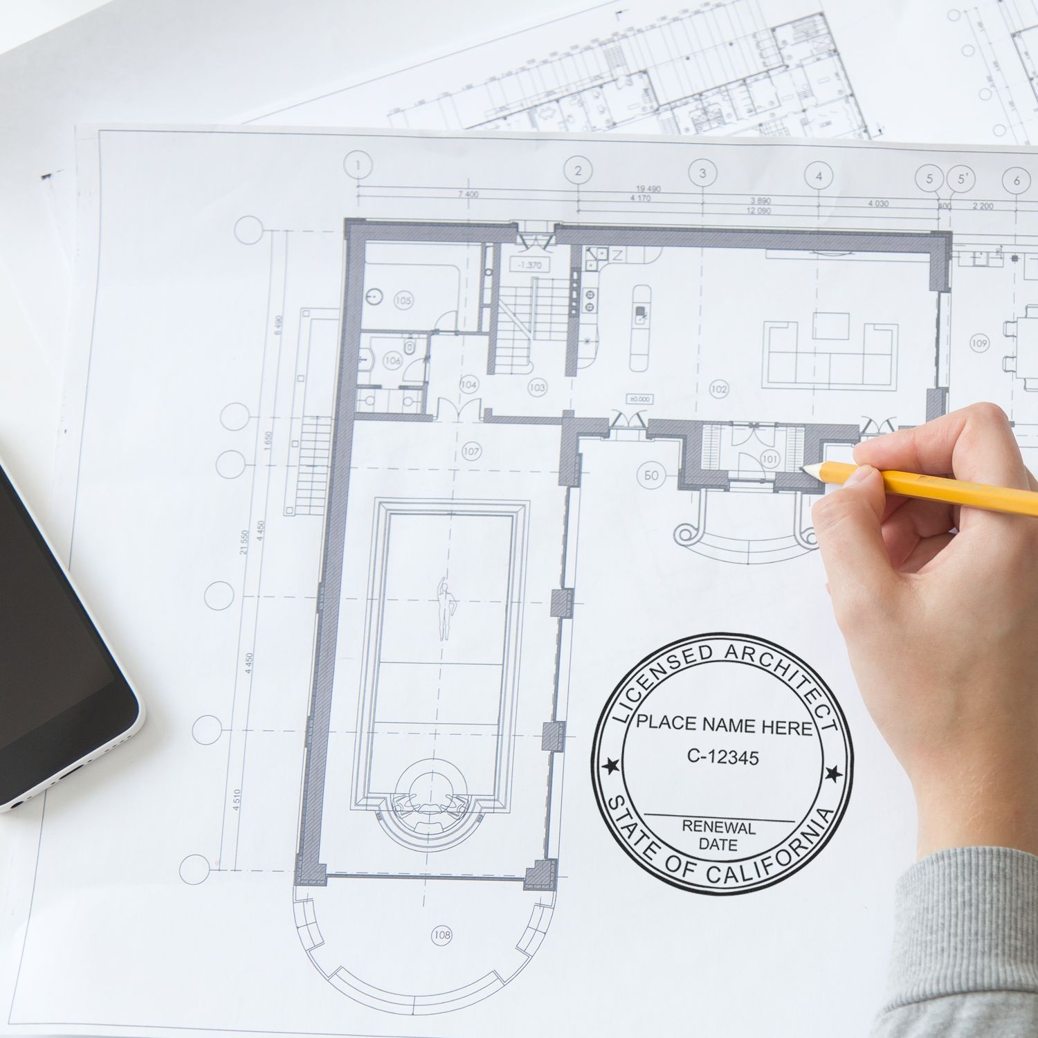 Person using Premium MaxLight Pre-Inked California Architectural Stamp on architectural blueprint with a pencil and smartphone nearby.