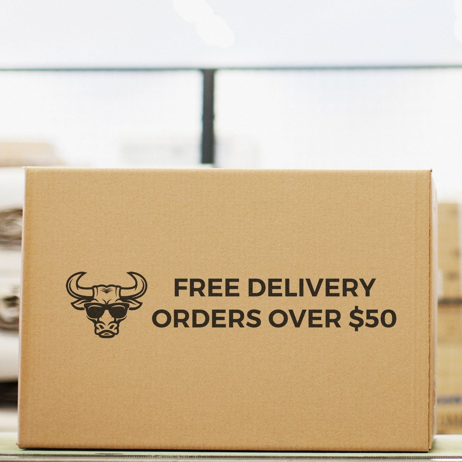 A cardboard box stamped with FREE DELIVERY ORDERS OVER $50 using a Custom Rubber Stamp Size 3 x 9, placed in a warehouse setting.