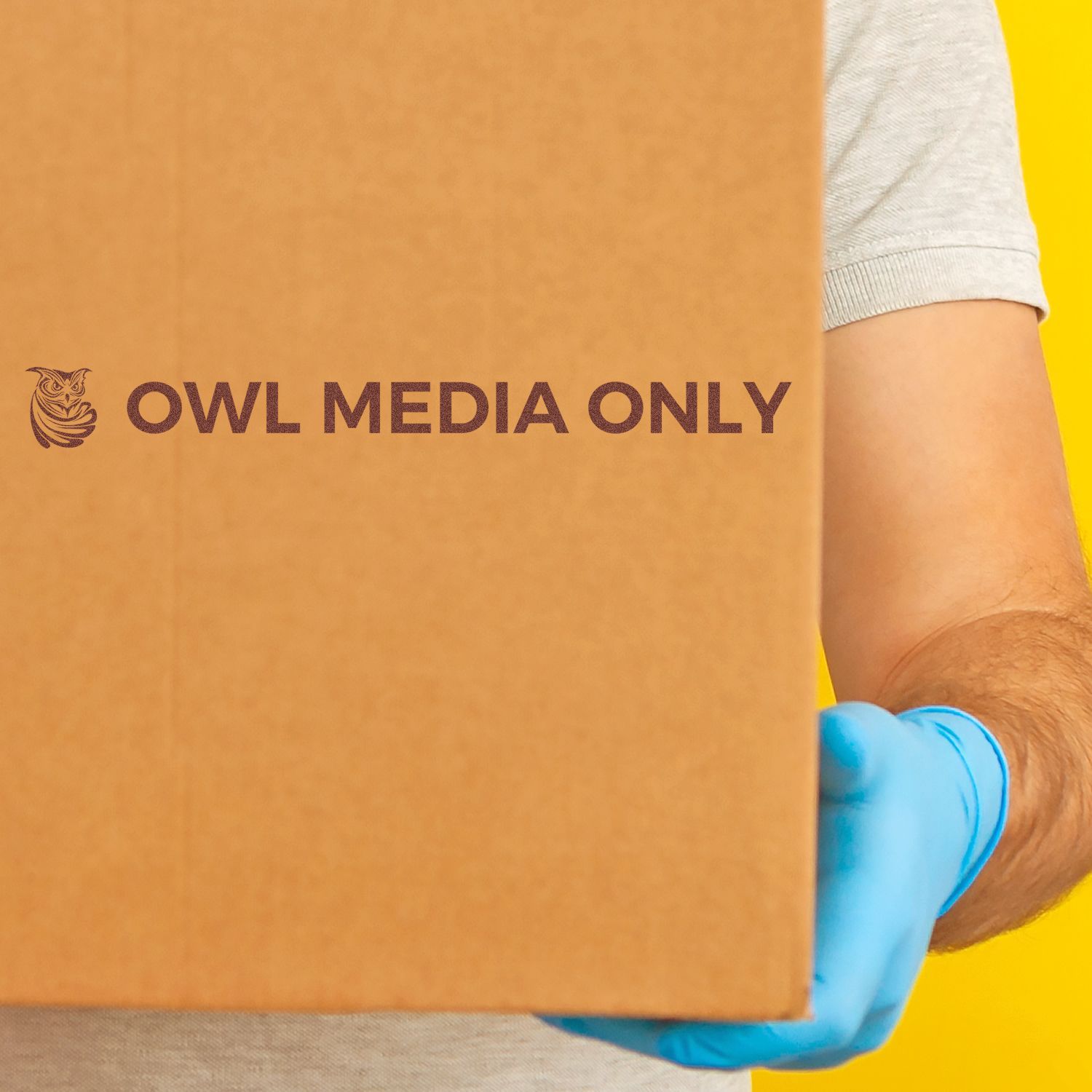 Person holding a cardboard box stamped with Owl Media Only using Custom Rubber Stamp Size 1-1/2 x 10, wearing blue gloves against a yellow background.