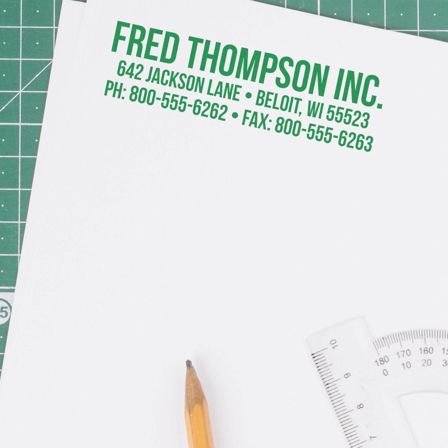 Custom Rubber Stamp Size 2 x 6 used to stamp green text on white paper, showing company details. Pencil and ruler nearby on a cutting mat.