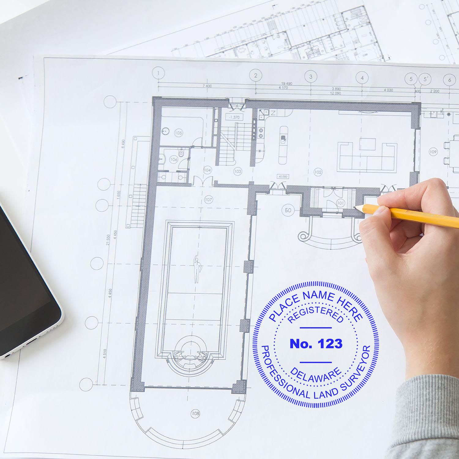 Person using Delaware Land Surveyor Seal Stamp, DE PLS Stamp on architectural blueprint with pencil and smartphone nearby.