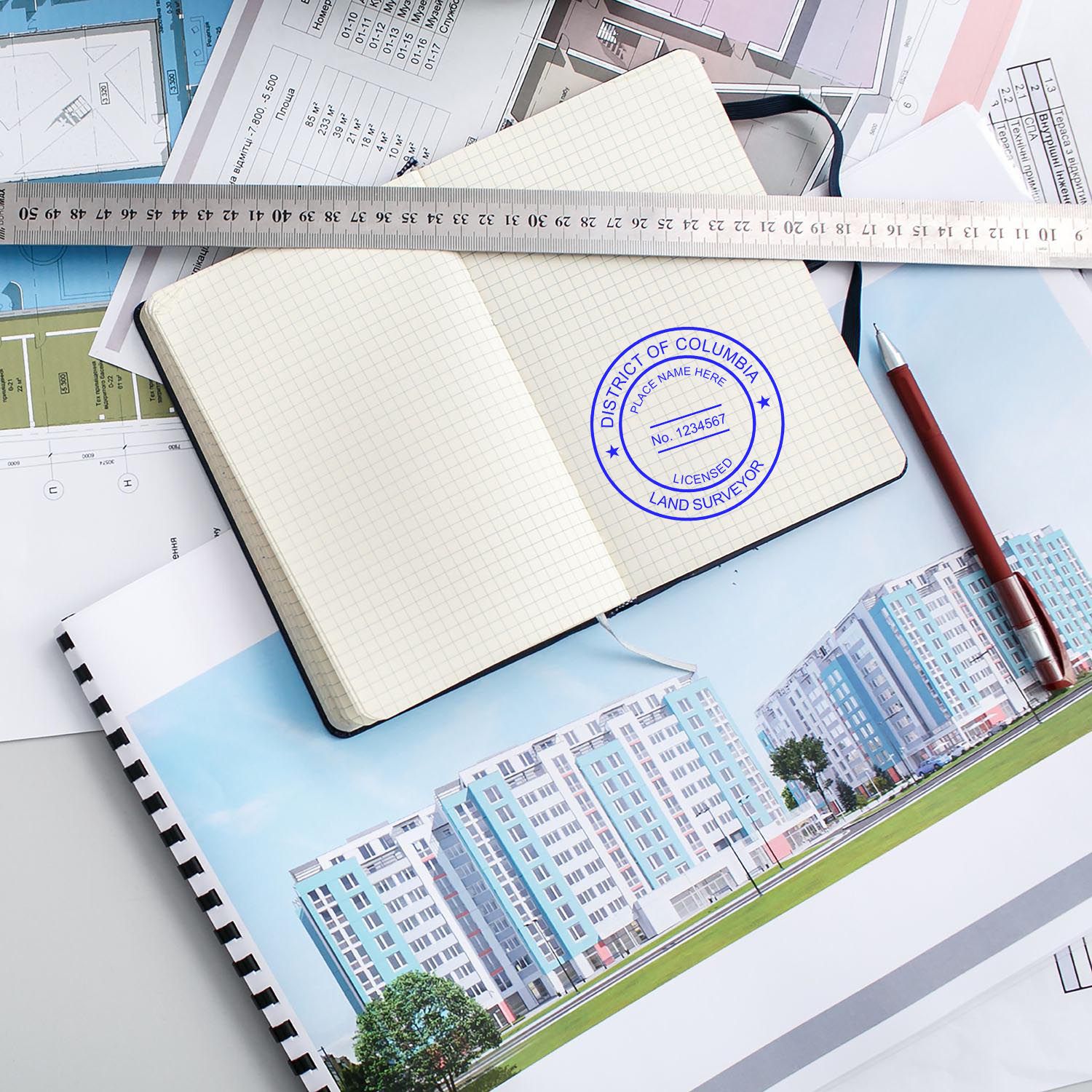 Self Inking District of Columbia Land Surveyor Stamp on an open notebook, surrounded by architectural plans, a ruler, and a pen.