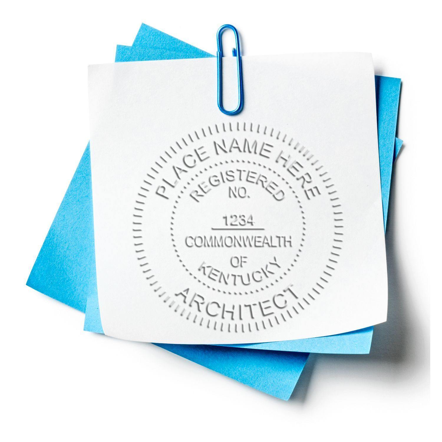 The image shows a white paper embossed with an architect seal, held by a blue paper clip on top of blue sheets. The product used is the Architect Long Reach Desk Seal Embosser.