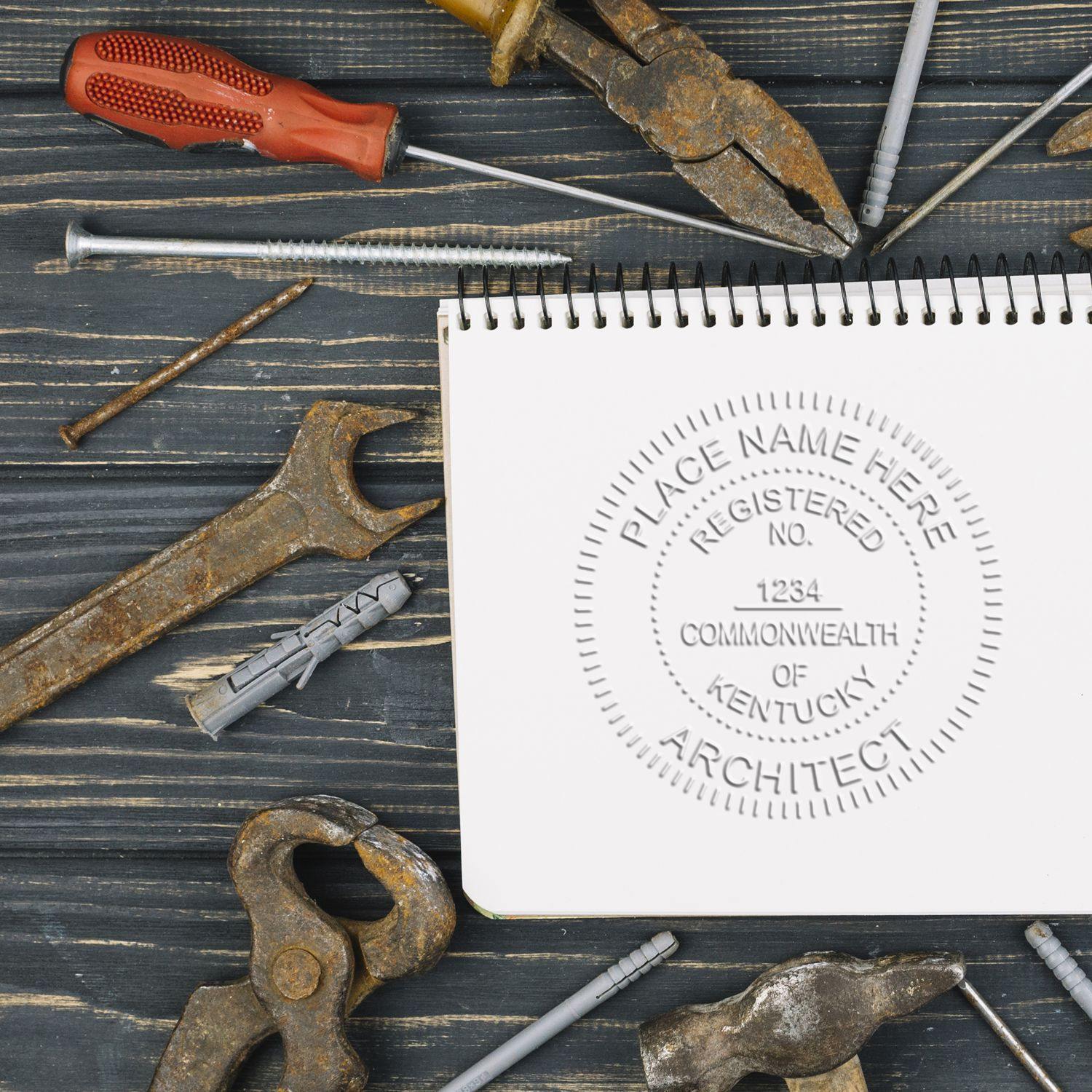 Architect Desk Seal Embosser imprint on a spiral notebook surrounded by various tools including a wrench, pliers, screwdriver, and nails on a wooden surface.