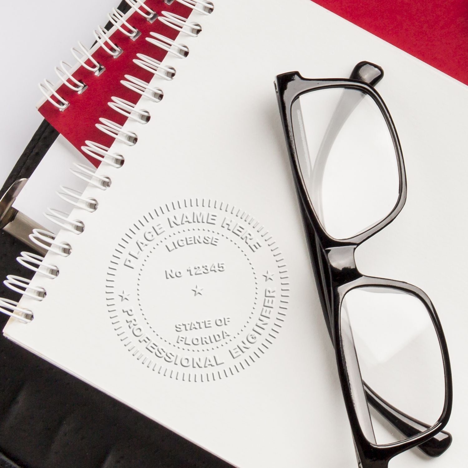 Professional Engineer Gold Gift Seal Embosser imprint on a white notebook page, with black-rimmed glasses and red and black notebooks in the background.