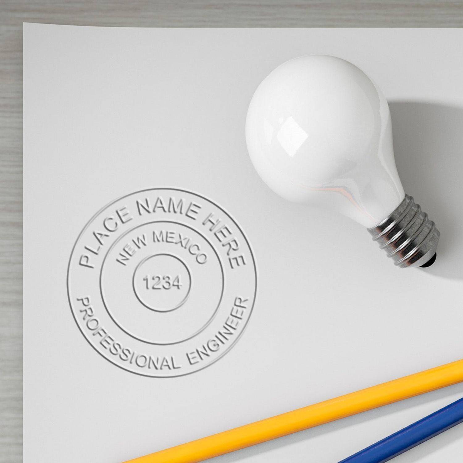 A close-up of a white paper embossed with a seal using the Professional Engineer Chrome Gift Seal Embosser, placed next to a light bulb and two pencils. The seal reads PLACE NAME HERE, NEW MEXICO, 1234, PROFESSIONAL ENGINEER.