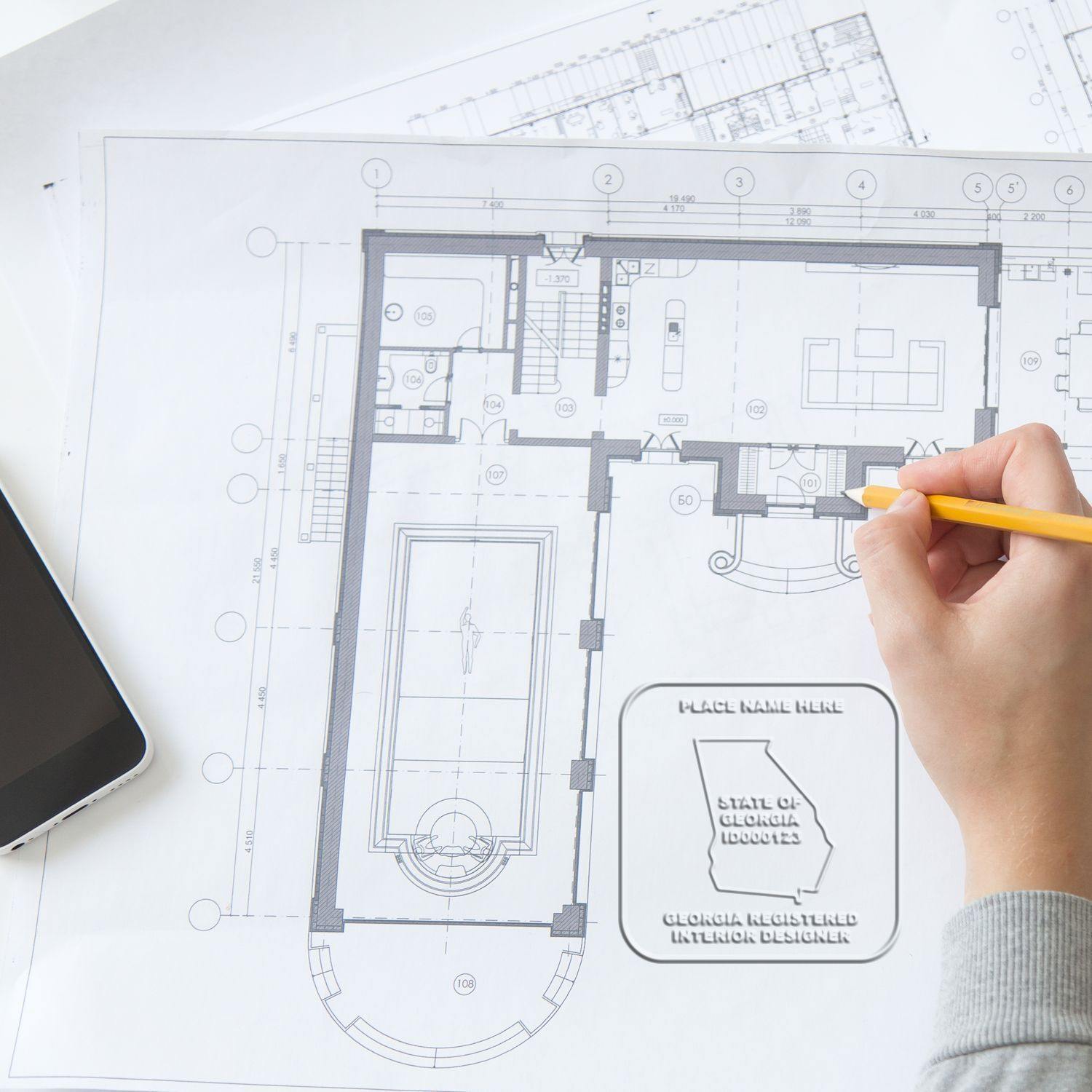 A hand using the Interior Designer Cast Iron Desk Seal Embosser on architectural blueprints, with a phone and pencil nearby.