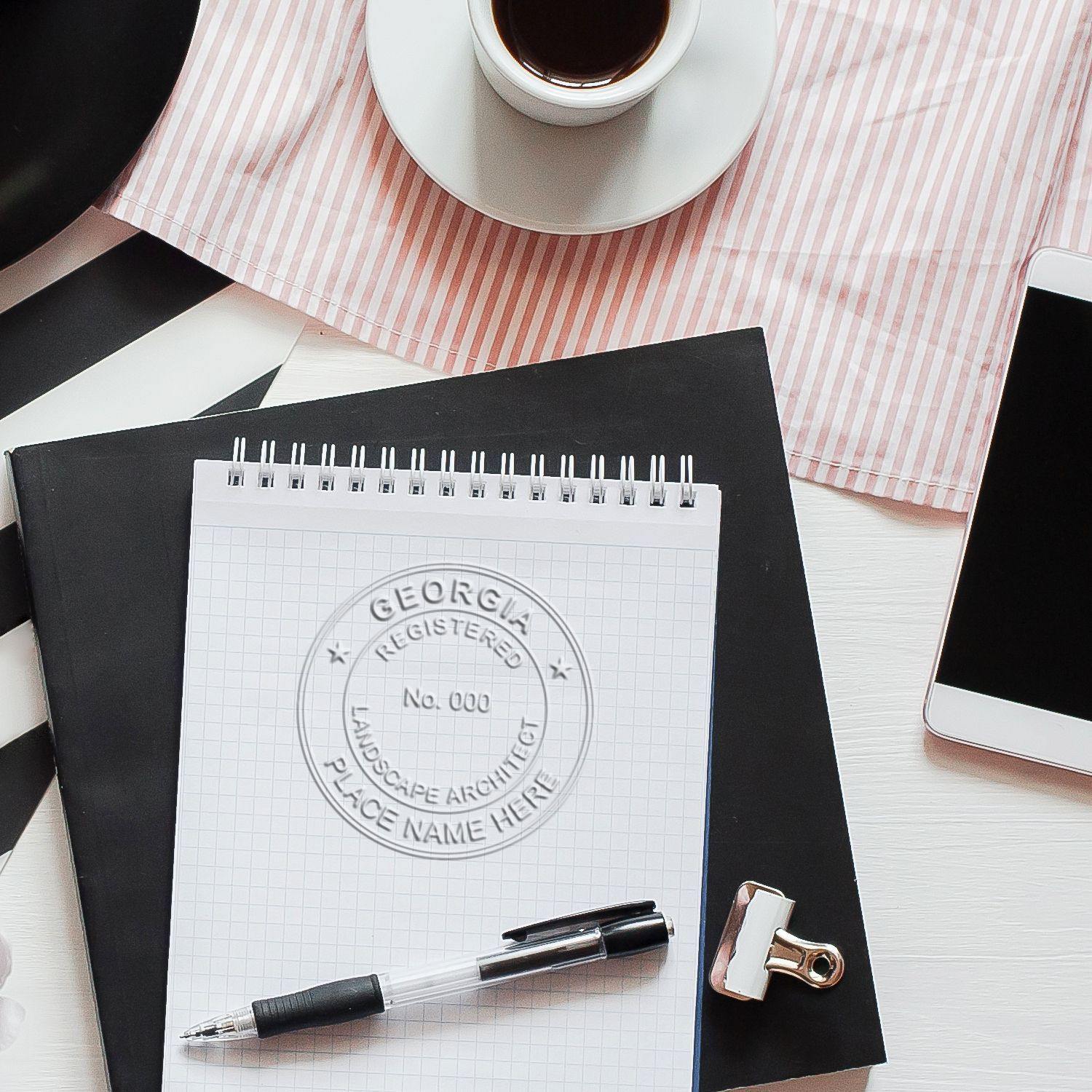 A Landscape Architect Desk Seal Embosser imprint on a notepad, surrounded by a pen, clipboard, coffee cup, and smartphone on a desk.