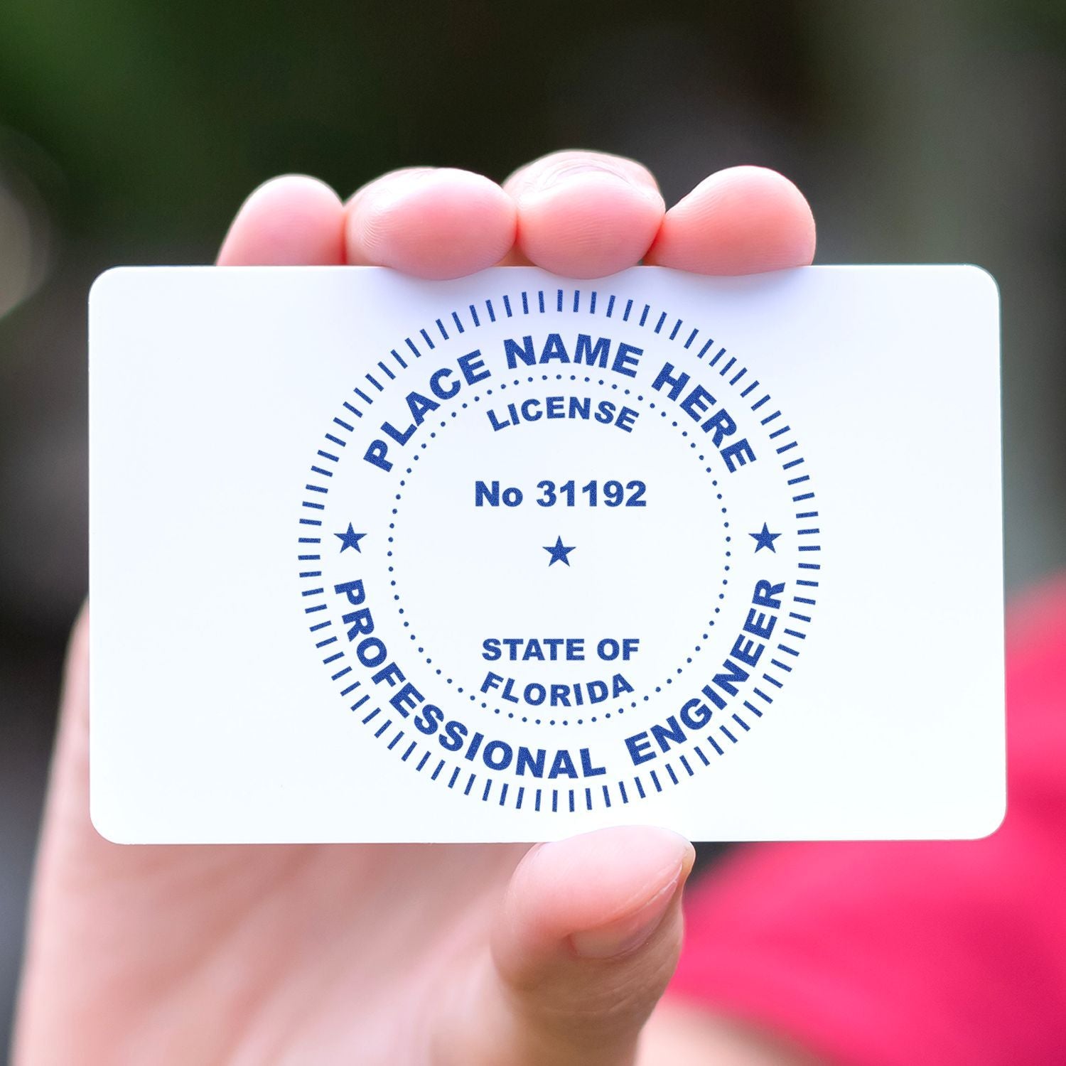 A hand holding a white card stamped with the Xstamper Professional Engineer Pre-Inked Stamp of Seal, displaying PLACE NAME HERE, LICENSE, No 31192, STATE OF FLORIDA, and PROFESSIONAL ENGINEER.