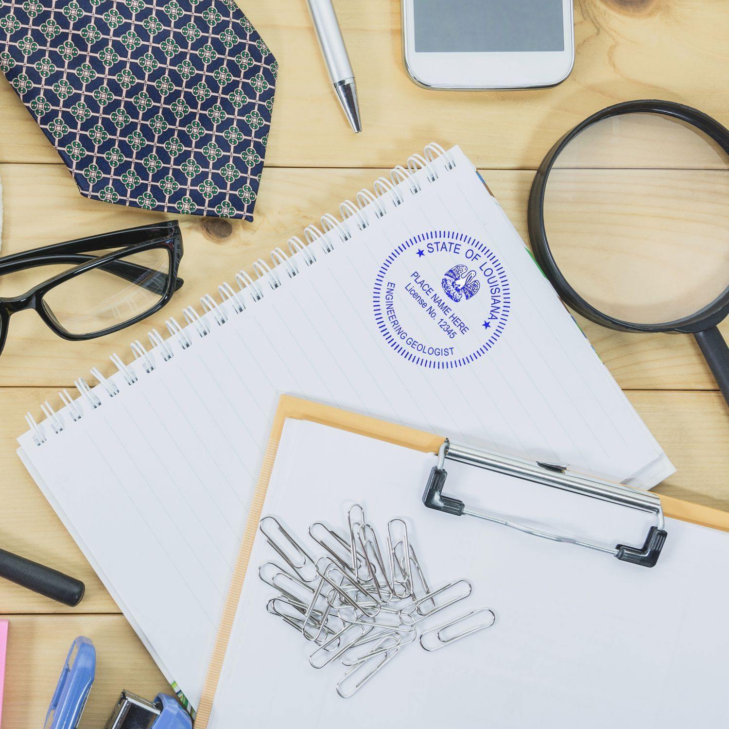 A desk with office supplies, including a notepad stamped with the Engineering Geologist MaxLight Pre-Inked Rubber Stamp of Seal, glasses, a tie, a magnifying glass, paper clips, and a smartphone.