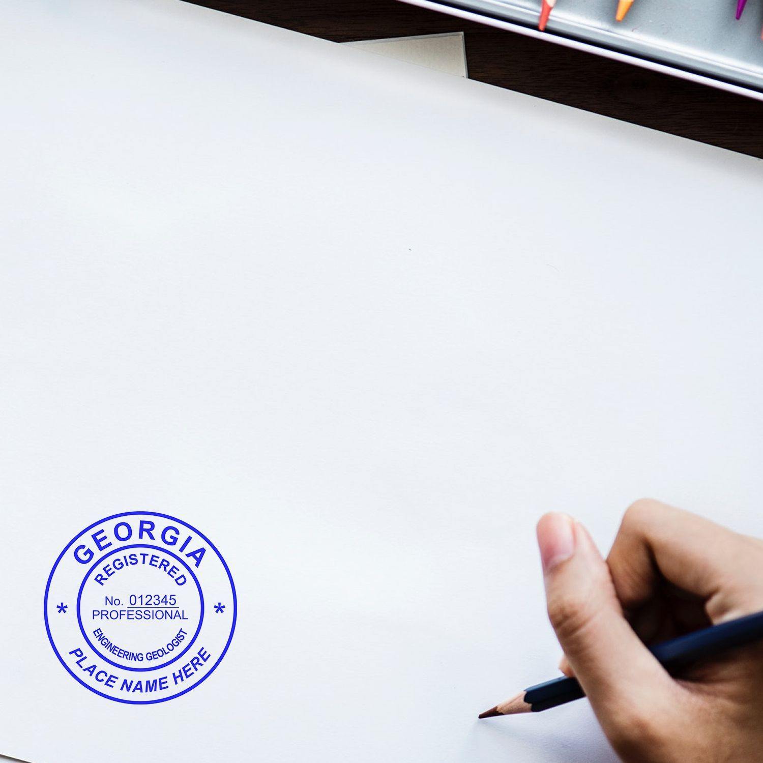 Hand holding pencil near a stamped paper with the Engineering Geologist MaxLight Pre-Inked Rubber Stamp of Seal, showing a blue circular seal with Georgia Registered Professional Engineering Geologist text.