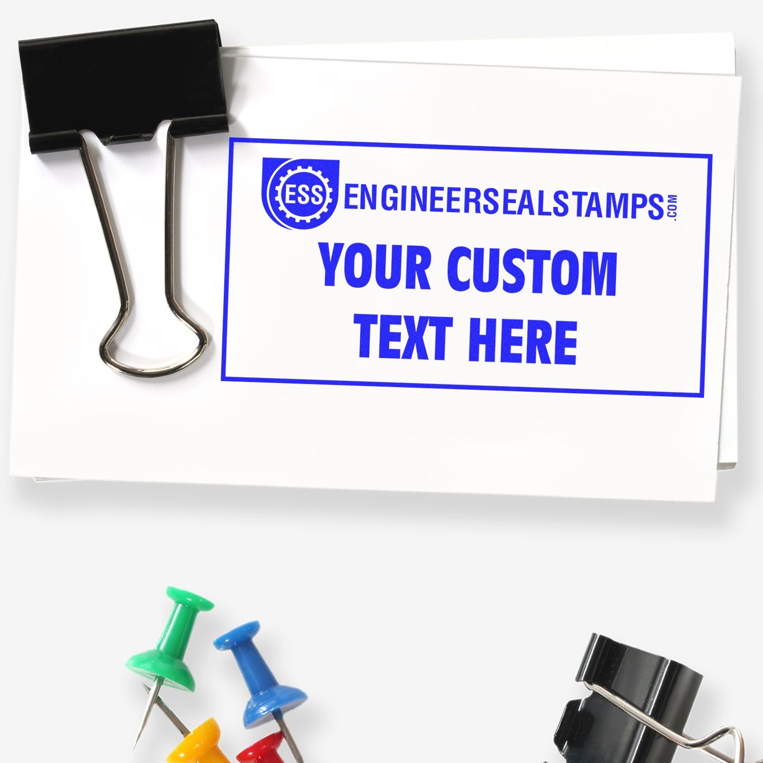 Custom Rubber Stamp Size 5 x 10 used on a white paper with YOUR CUSTOM TEXT HERE in blue, next to a black binder clip and colorful push pins.