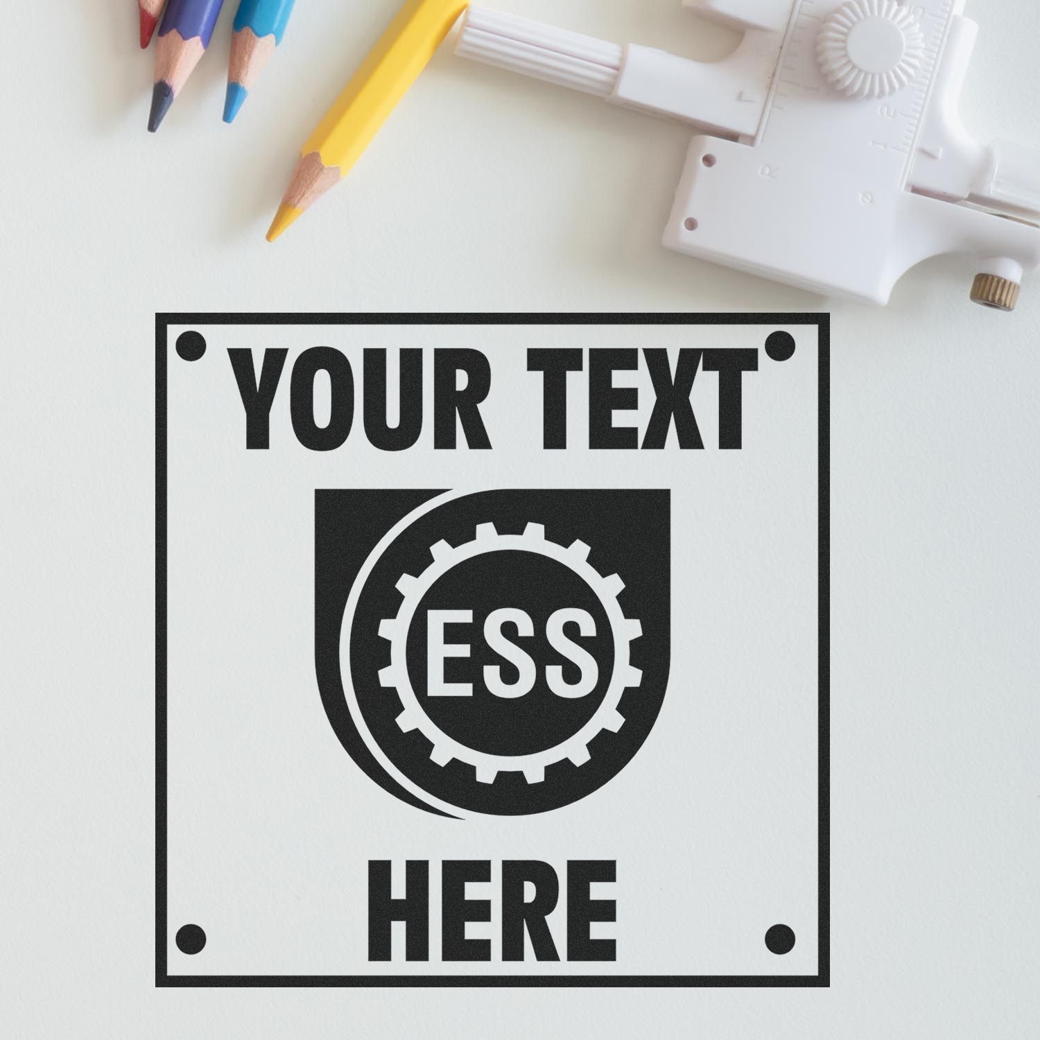 Custom Rubber Stamp Size 6 x 6 with YOUR TEXT HERE and ESS logo, placed on a white surface with pencils and a tool nearby.