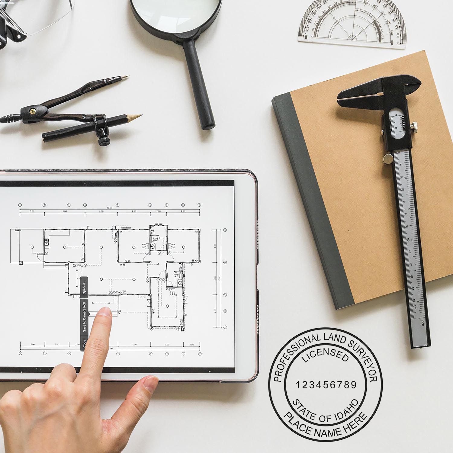 Self Inking Idaho Land Surveyor Stamp in use on a desk with architectural tools, a tablet displaying a blueprint, and a notebook.