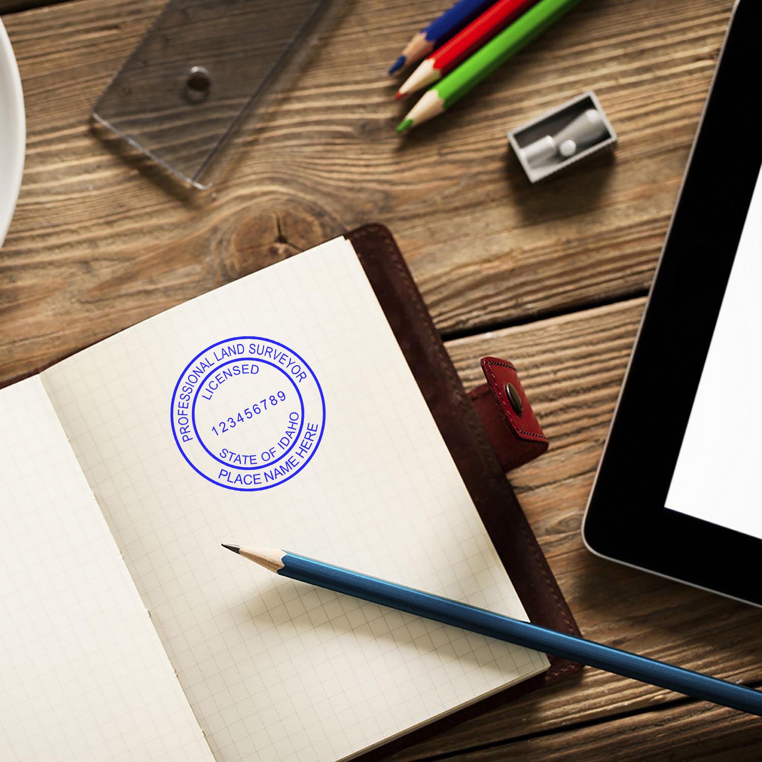 Self Inking Idaho Land Surveyor Stamp on an open notebook with a pencil, colored pencils, sharpener, and tablet on a wooden desk.