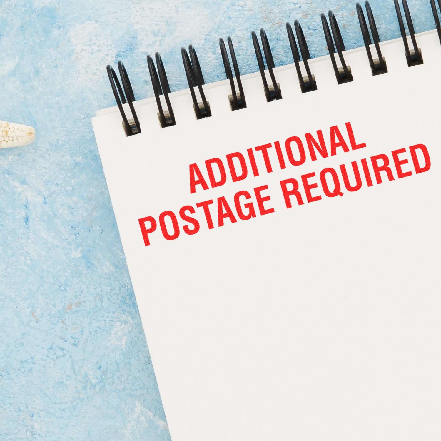 A spiral notebook with a red Additional Postage Required rubber stamp imprint on a light blue textured background.