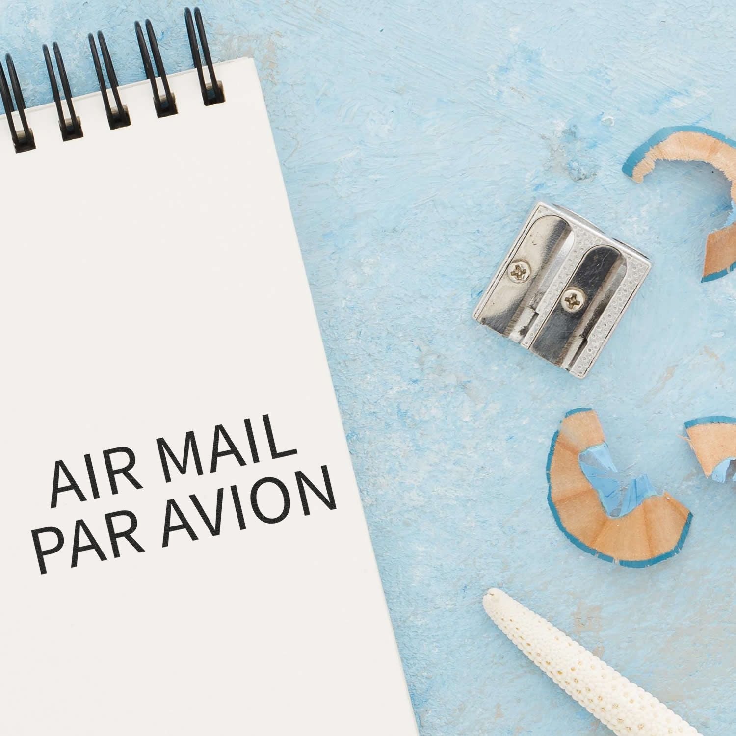 A notebook with AIR MAIL PAR AVION text, a pencil sharpener, and pencil shavings on a light blue surface.