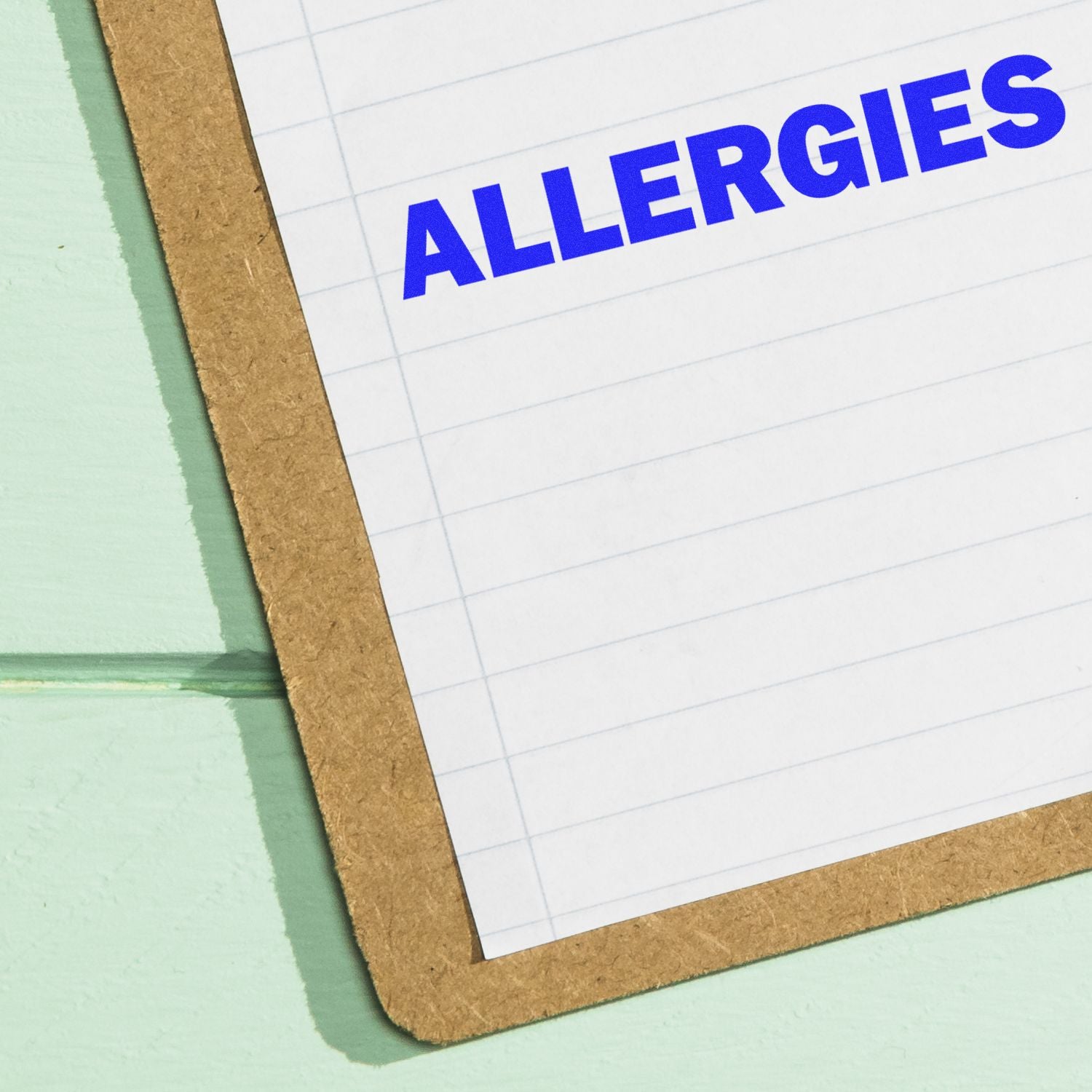 Allergies rubber stamp in blue ink on a lined paper attached to a clipboard, placed on a light green surface.