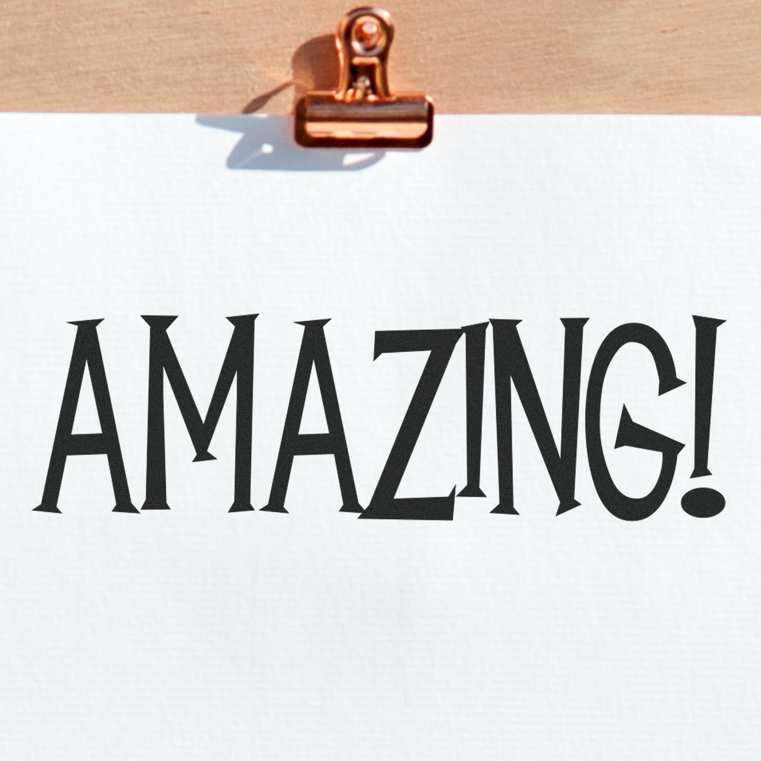 Amazing Rubber Stamp imprint on white paper clipped to a wooden board, showcasing bold, stylized letters spelling 'AMAZING!'