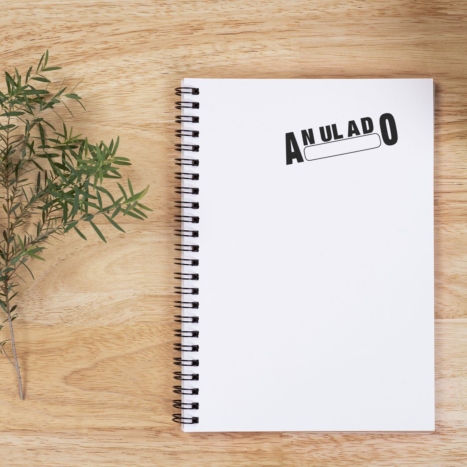 Large Anulado Rubber Stamp marking ANULADO on an open spiral notebook, placed on a wooden surface next to a small green plant.