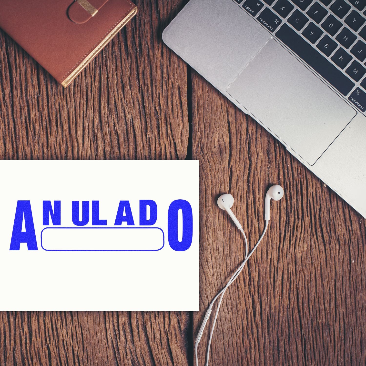 Large Self Inking Anulado Stamp in blue ink on paper, placed on a wooden desk next to a laptop, notebook, and earphones.