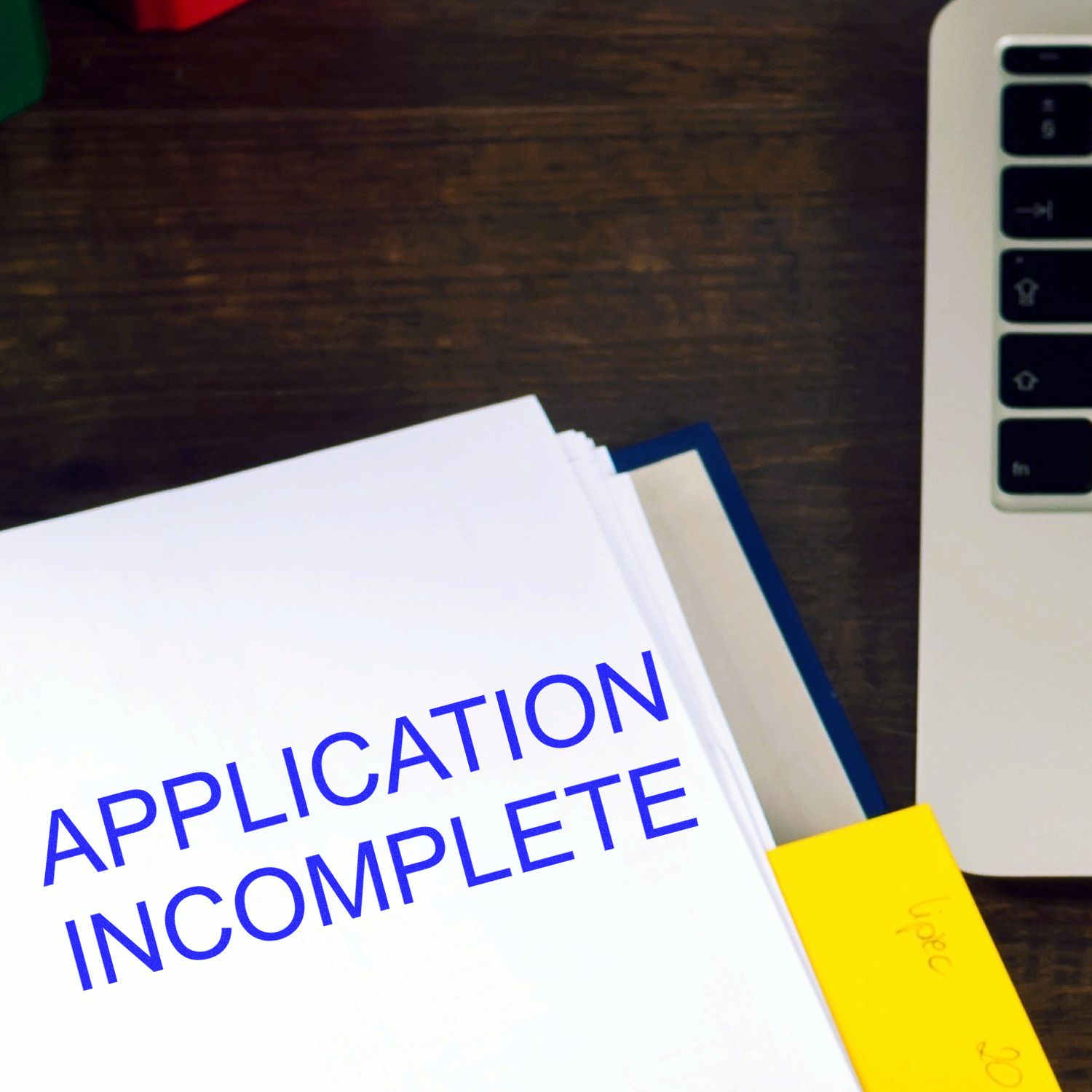 Application Incomplete rubber stamp on a stack of papers next to a laptop on a wooden desk.