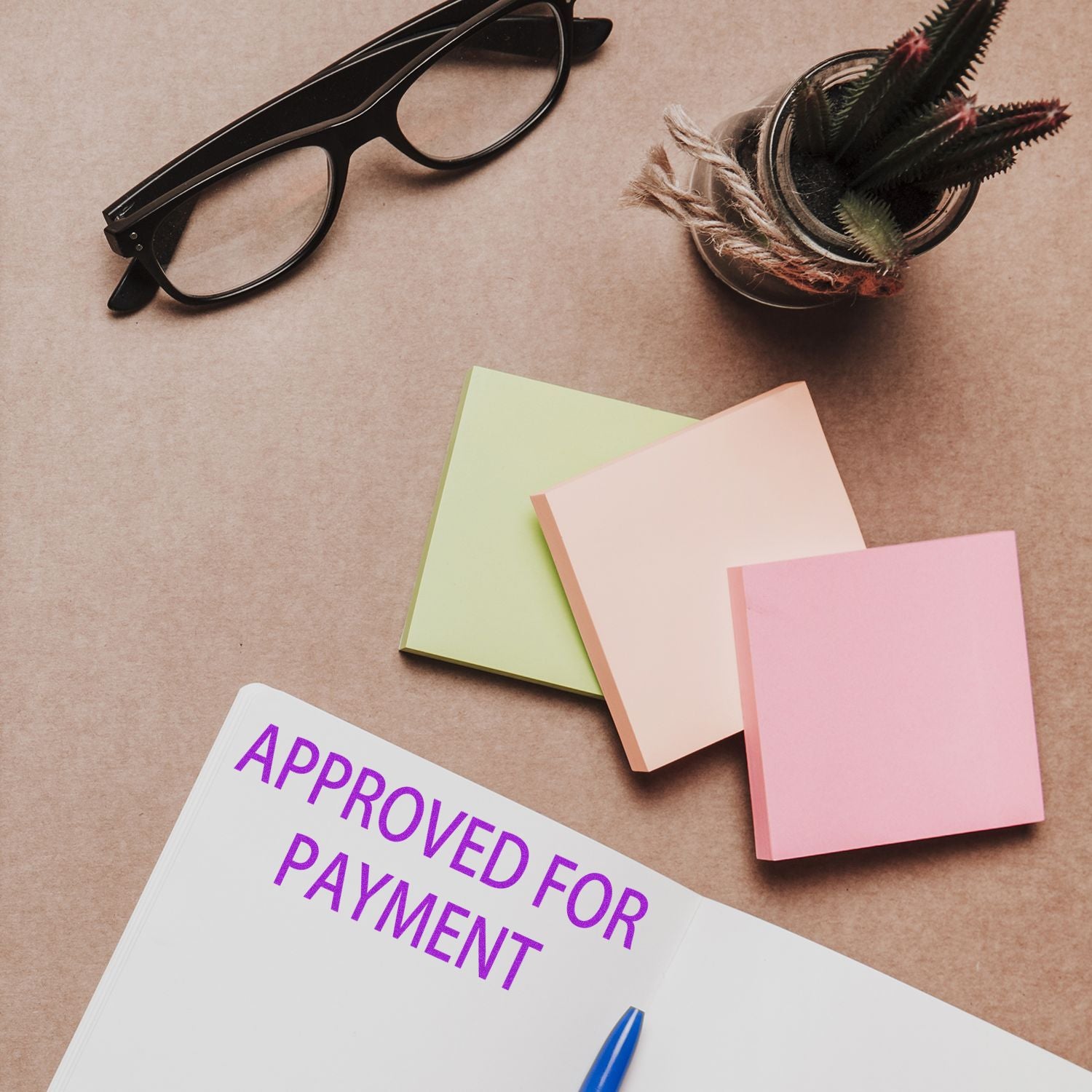 Large Approved For Payment Rubber Stamp used on a document, with sticky notes, glasses, and a plant on a desk.