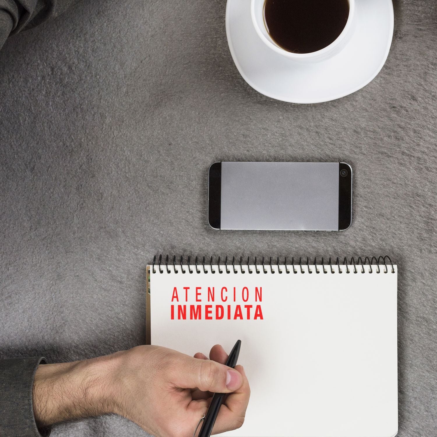 A person holding a pen next to a notebook stamped with Atencion Inmediata Rubber Stamp, a smartphone, and a cup of coffee.