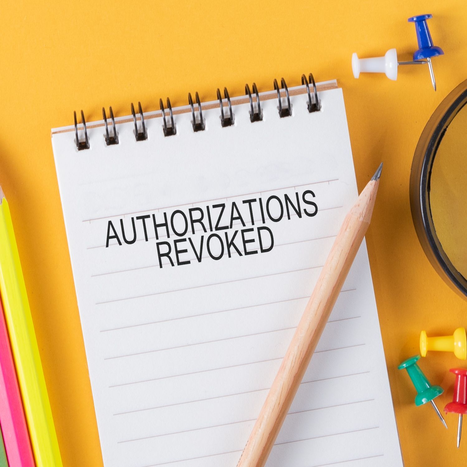 A notepad with AUTHORIZATIONS REVOKED written, surrounded by a pencil, rubber stamp, and colorful push pins on a yellow background.