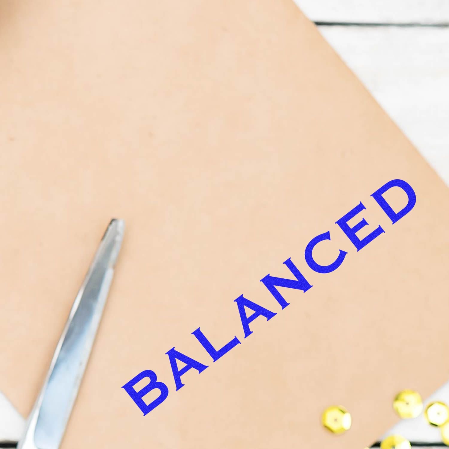 A stamped paper with the word BALANCED in blue ink using the Self Inking Balanced Stamp, next to a pair of scissors and sequins.