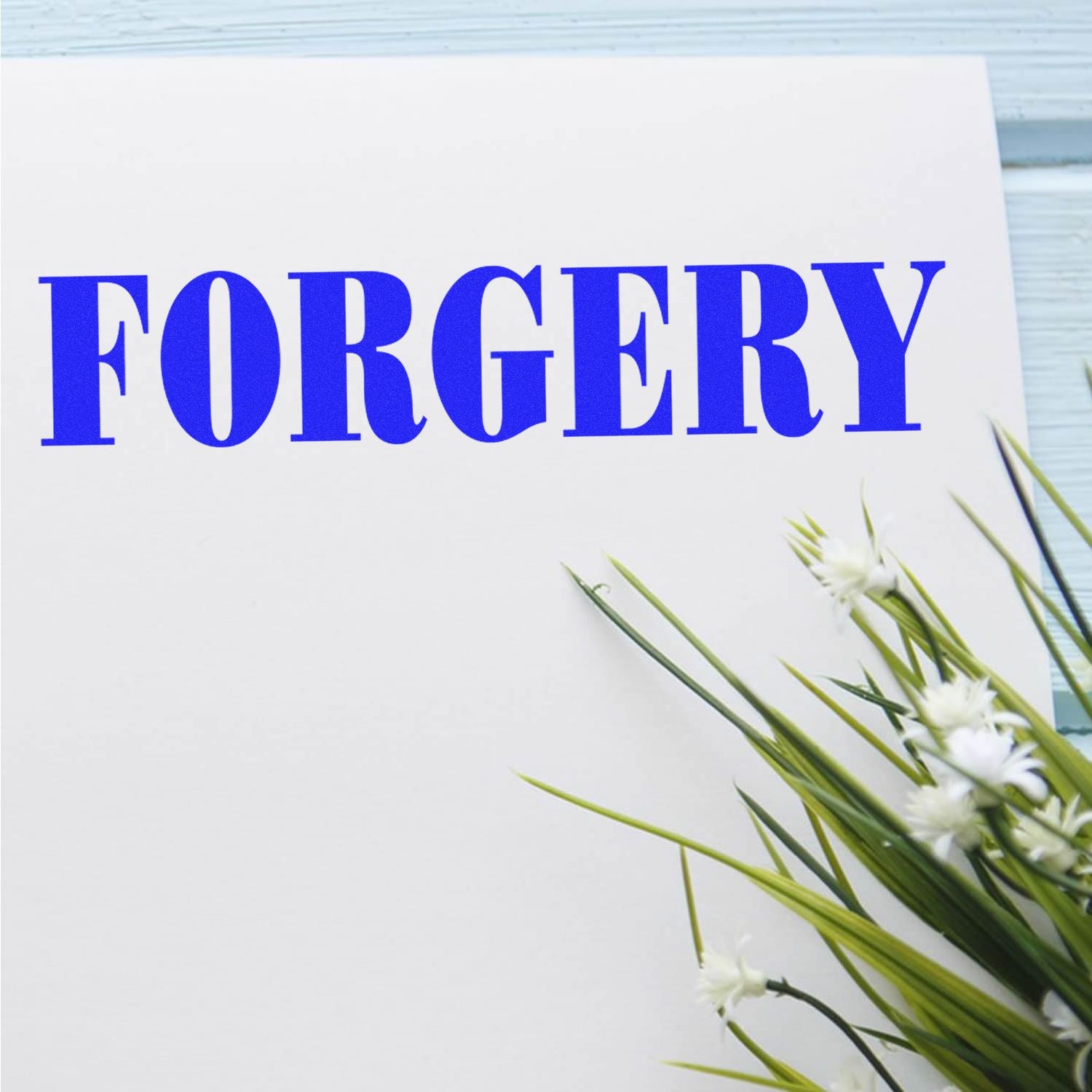 Paper stamped with 'FORGERY' in blue ink using a Slim Pre-Inked Forgery Stamp, next to green and white flowers on a light blue surface.