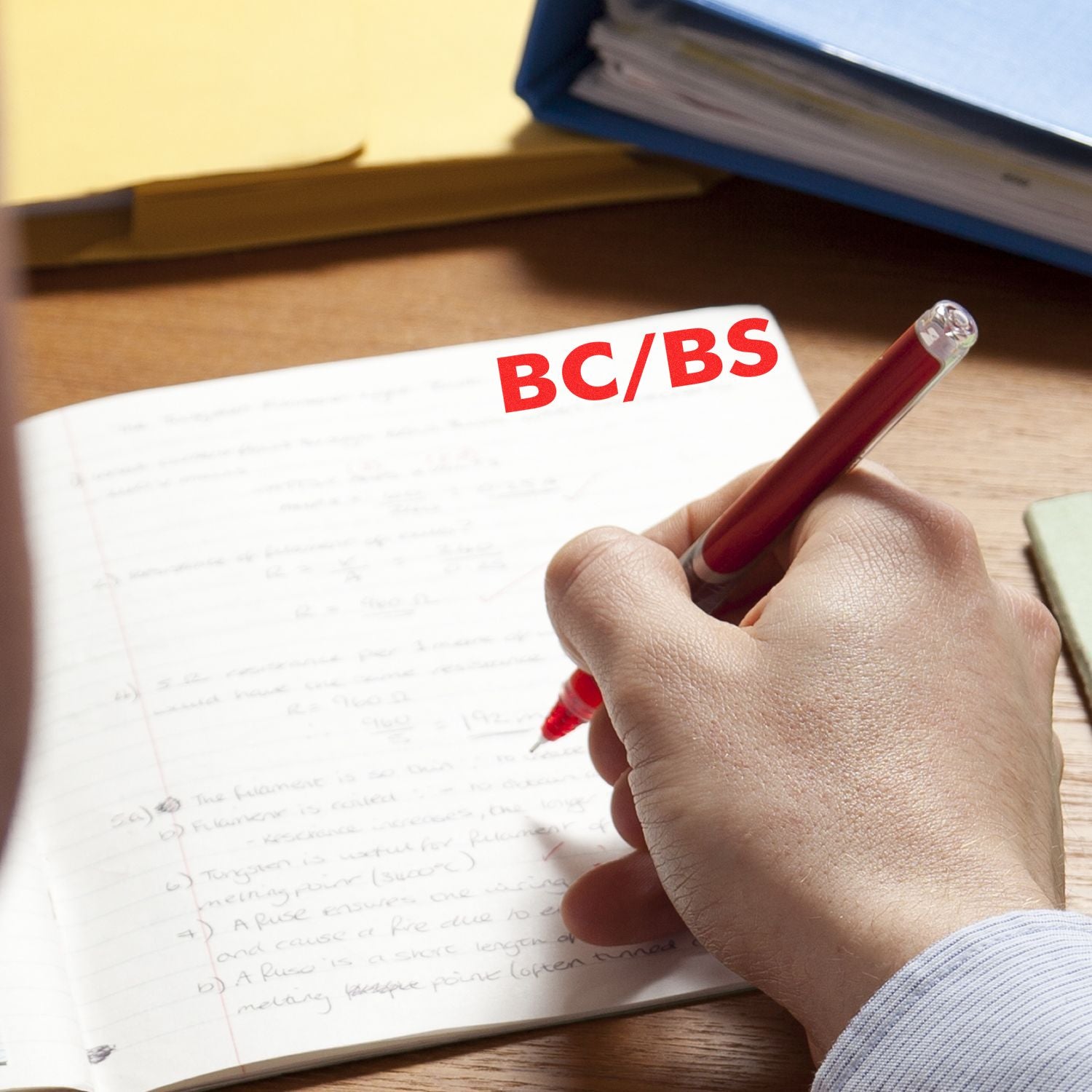 Hand using a red pen to write in a notebook with the Self Inking BC/BS Stamp imprint visible on the page, surrounded by office supplies.