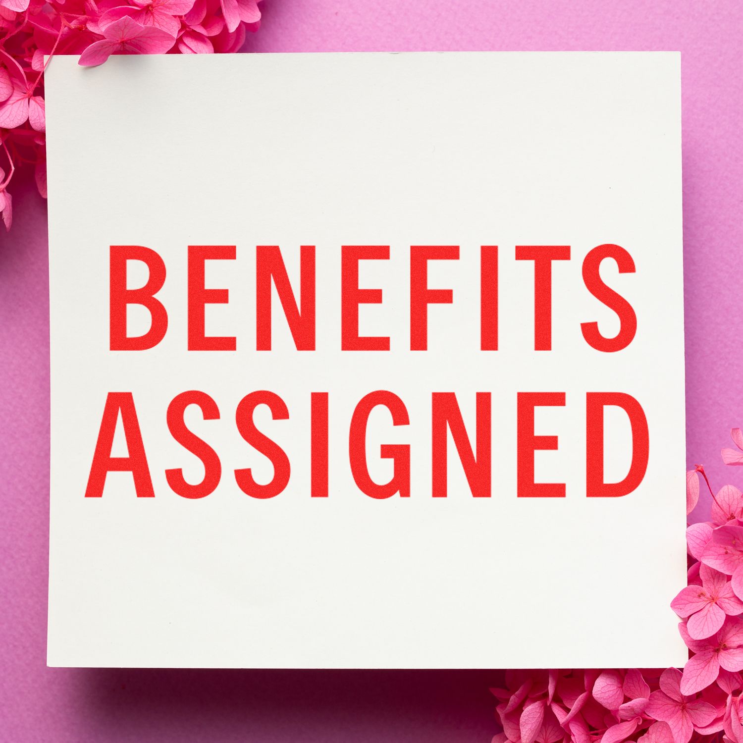 Slim Pre-Inked Benefits Assigned Stamp in red ink on white paper, surrounded by pink flowers on a pink background.