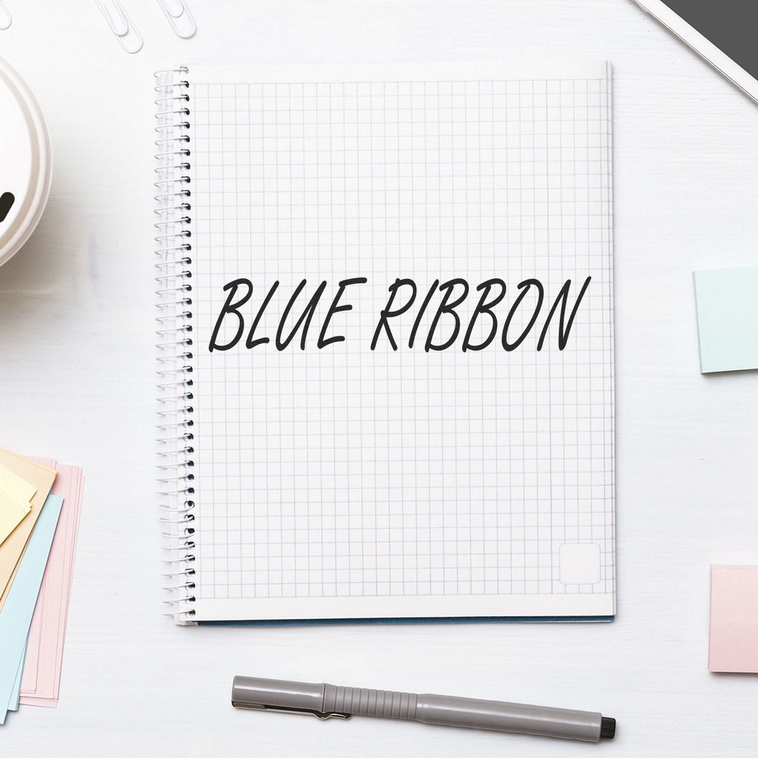 Large Self Inking Blue Ribbon Stamp in use on a spiral notebook with BLUE RIBBON text, surrounded by stationery items on a white desk.