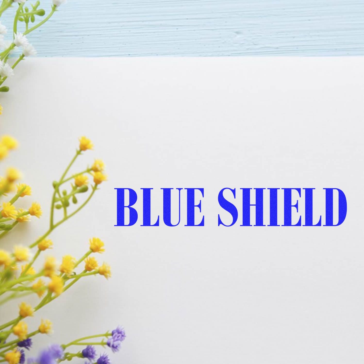 Large Blue Shield Rubber Stamp imprint on white paper with yellow and purple flowers in the corner, against a light blue background.