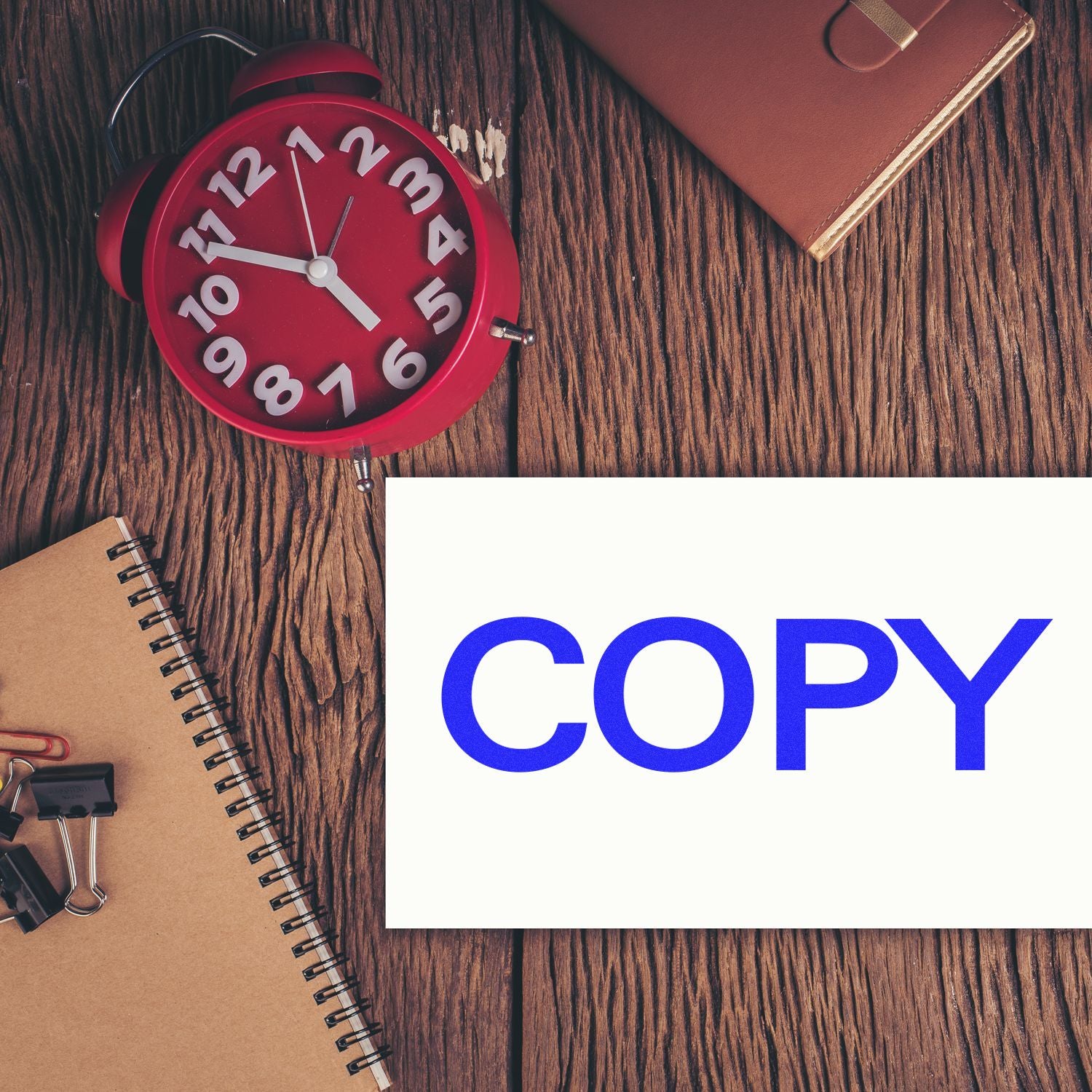 Large Self Inking Bold Copy Stamp in use on a white paper, placed on a wooden desk with a red clock, notebook, and office supplies.