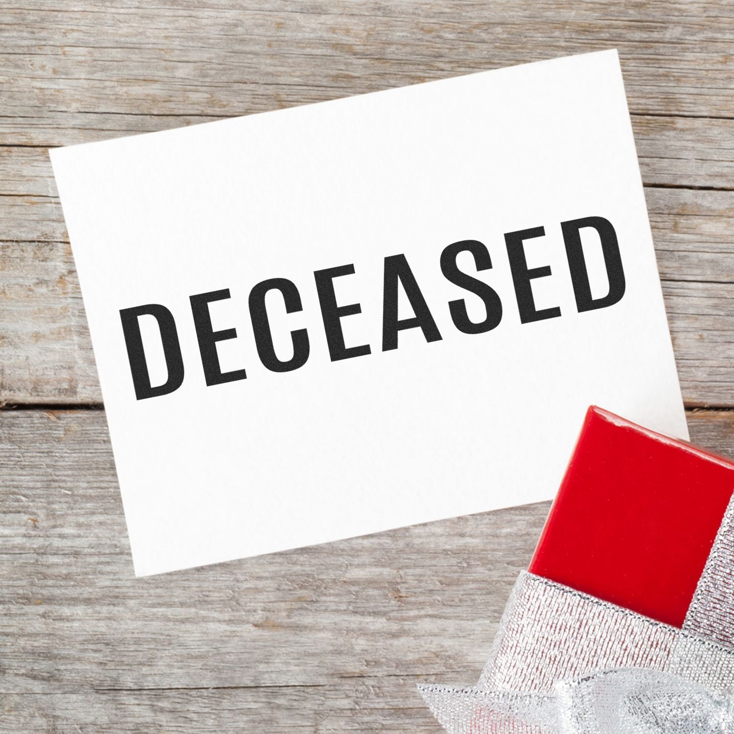 Bold Deceased Rubber Stamp on a white card placed on a wooden surface, with a red and silver gift box in the corner.