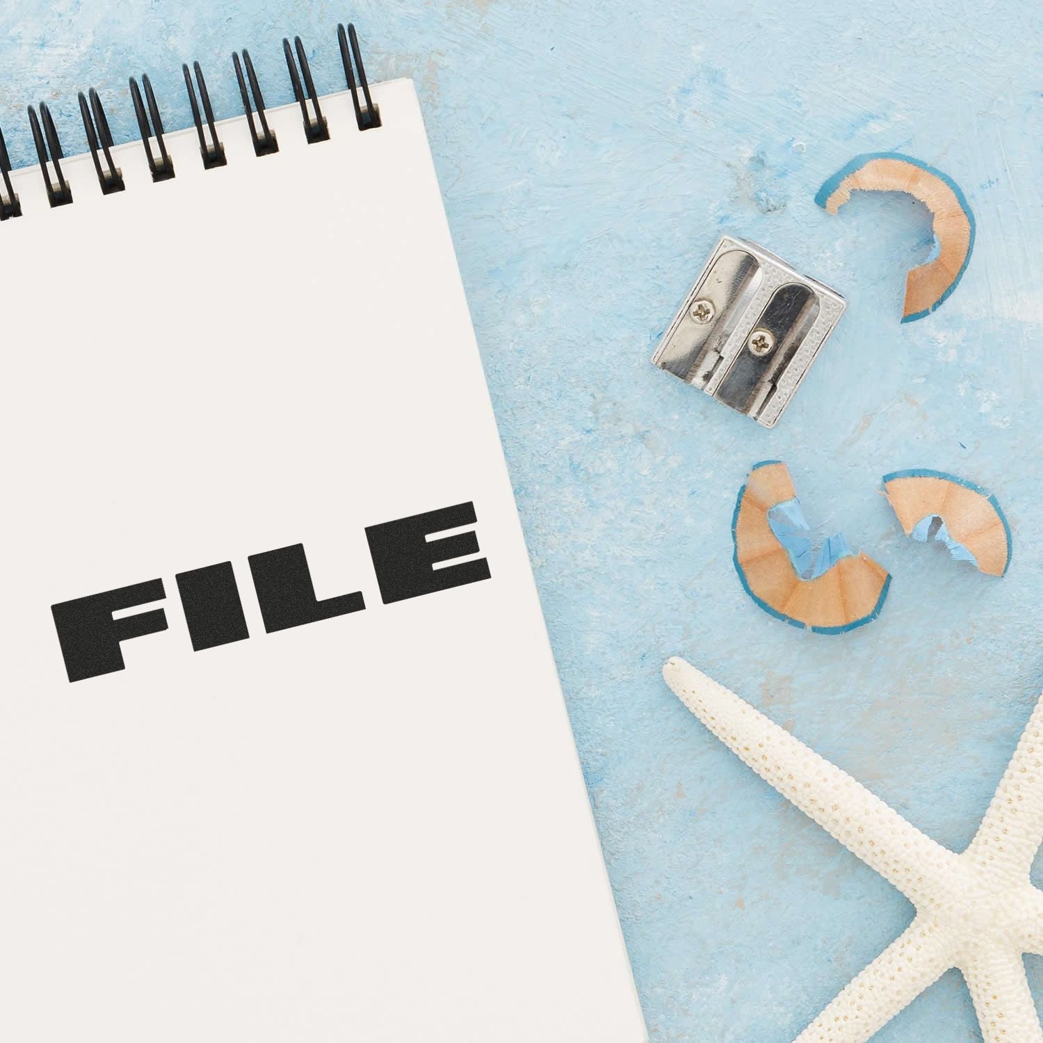 Large Self Inking Bold Font File Stamp used on a white notepad, placed on a blue surface with a pencil sharpener, shavings, and a starfish.