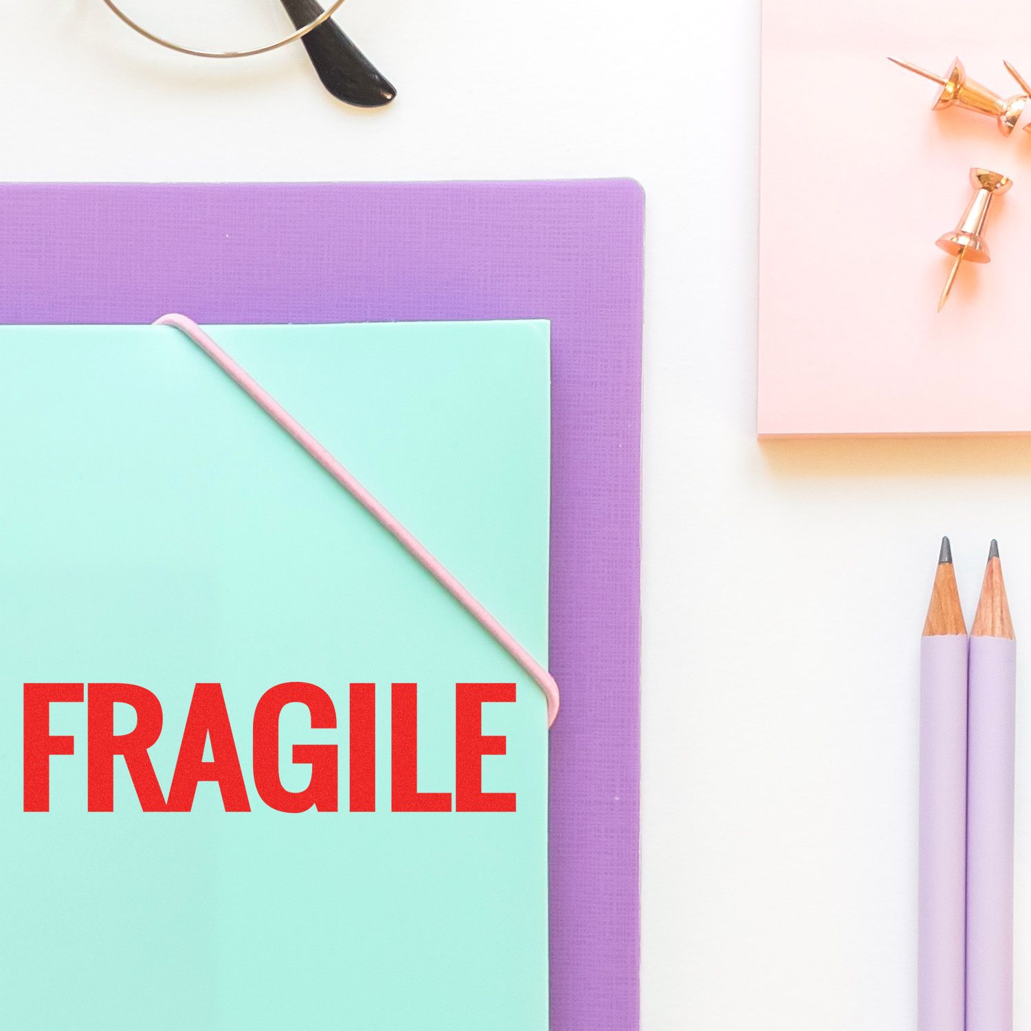 Bold Fragile Rubber Stamp in use on a mint green envelope, surrounded by office supplies including pencils, push pins, and a purple folder.