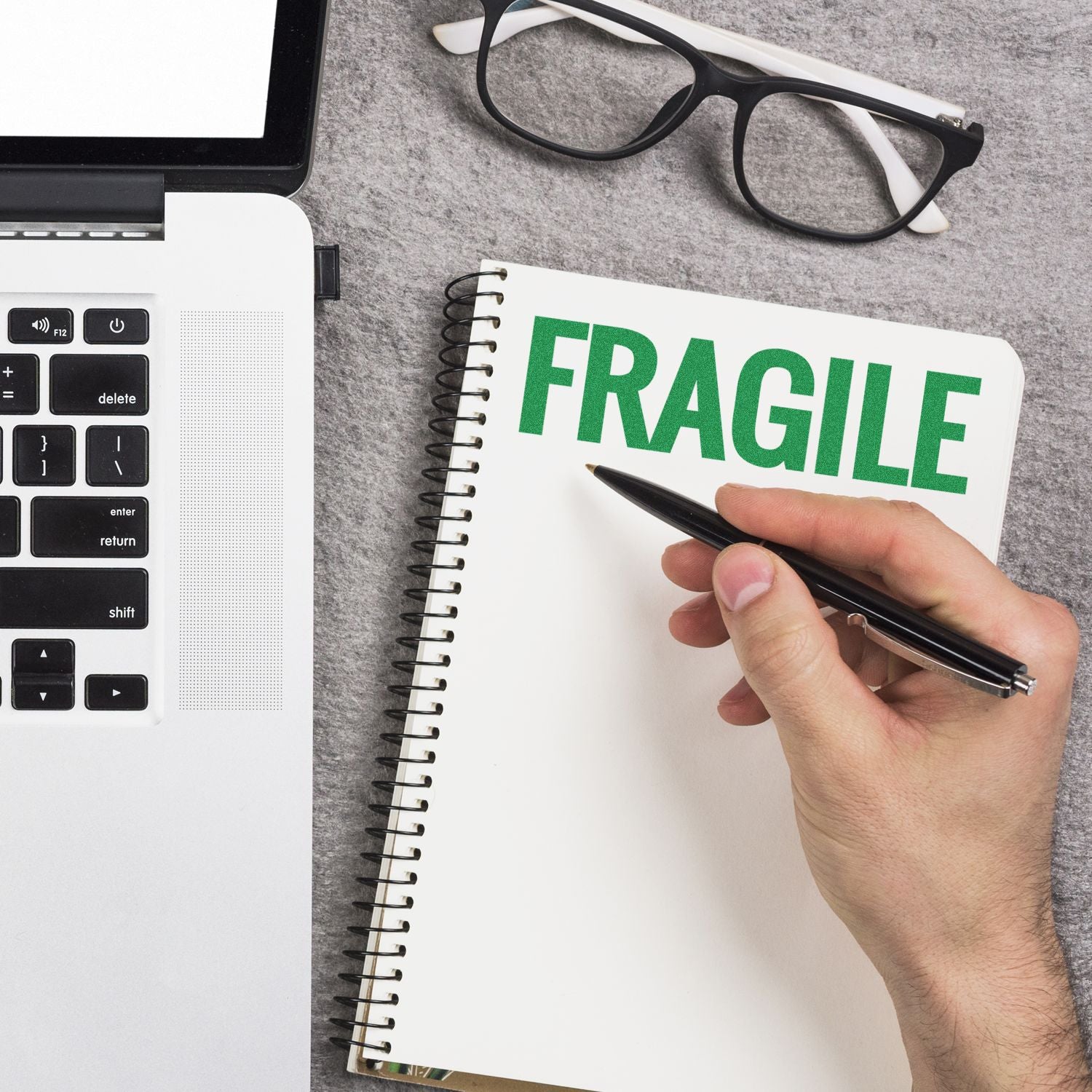 Hand holding a pen next to a notebook stamped with FRAGILE using the Large Bold Fragile Pre-Inked Stamp, placed beside a laptop and glasses.