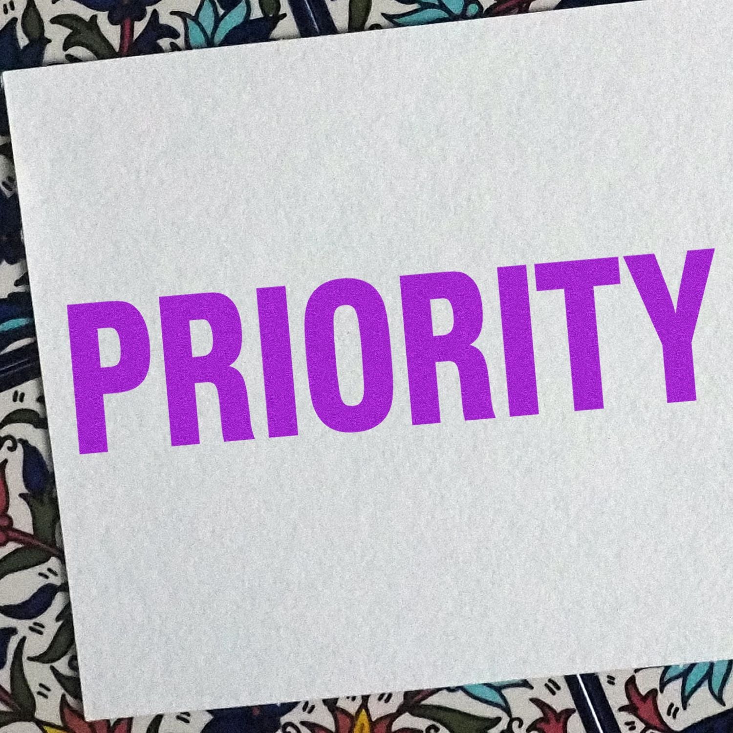 Bold Priority Rubber Stamp impression in purple ink on white paper, placed on a colorful patterned background.