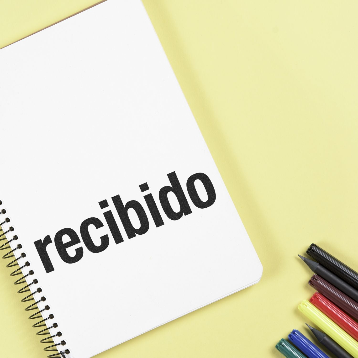 Bold Recibido Rubber Stamp used on a white spiral notebook, with colorful pens placed beside it on a yellow background.